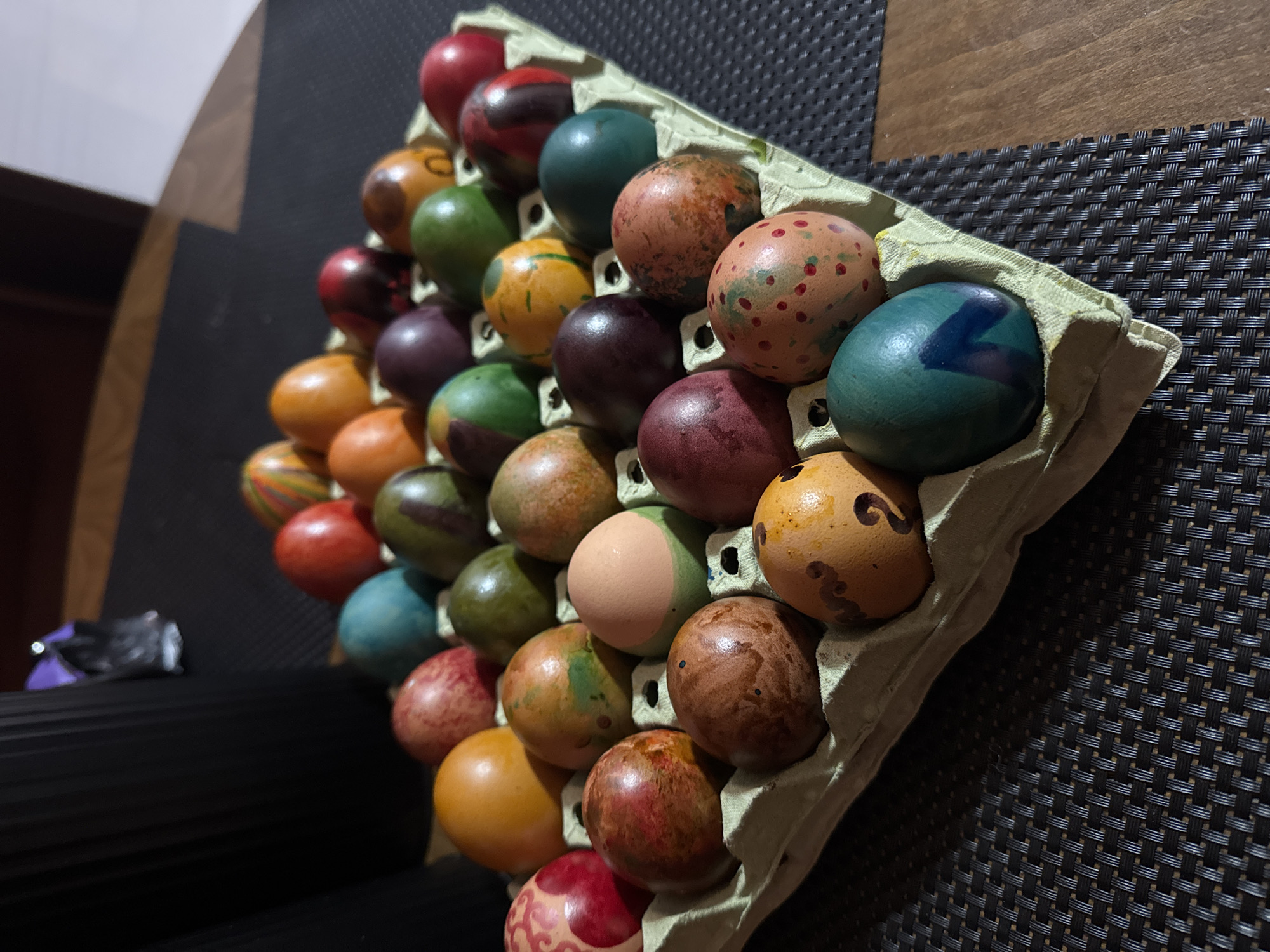 The image shows a 5x7 cardboard egg carton filled with painted Easter eggs. The eggs are decorated in a variety of colors and patterns. The carton sits on a dark, woven surface. The location and city cannot be determined from the visual information provided.