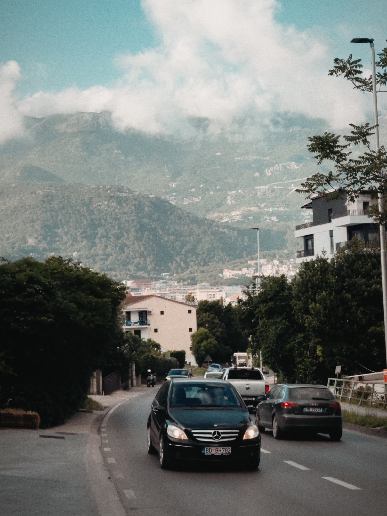 The image shows a street in Budva, Montenegro. The street curves gently uphill. In the foreground, a black Mercedes-Benz with license plate "BD BH792" is driving towards the camera, followed by a dark gray Audi with license plate "AB 962 DT". A white pickup truck is further behind. Trees line the street on both sides. A light-colored building is visible to the left of the road. Behind the building and through the trees, other buildings can be seen closer to the foreground. In the background, mountains are shrouded in clouds under a partly cloudy sky. A modern white building with balconies is to the right of the road, with a tall light pole next to it.
