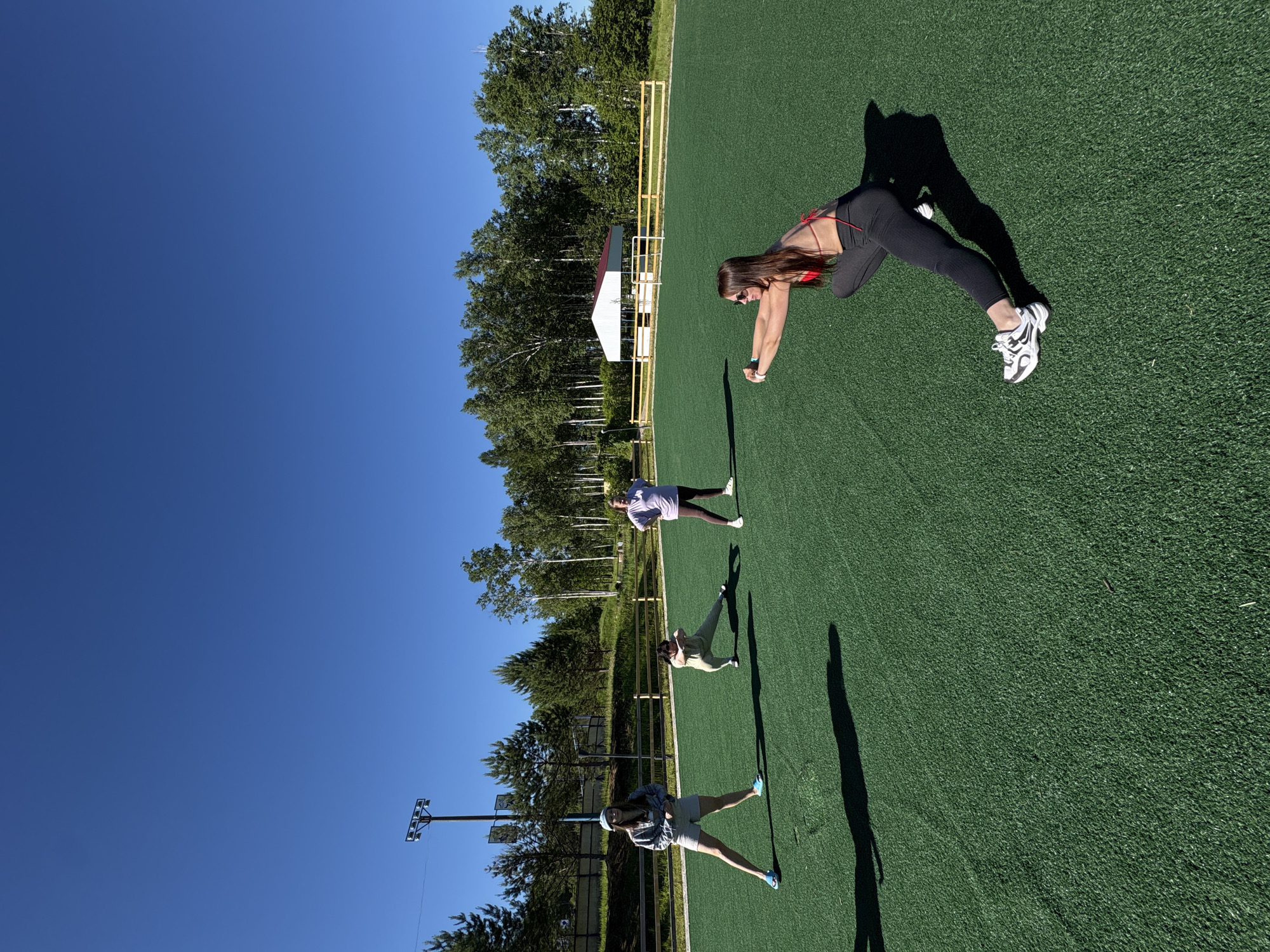 Four individuals are engaged in a group fitness session on an artificial turf field under a clear blue sky. The location is an outdoor sports field bordered by a wooden fence and a line of trees. In the background, a white shelter with a red roof and a tall lighting tower are visible.
From left to right:
The first person on the left is wearing a light blue cap, a gray hooded sweatshirt, and light-colored shorts, with blue sandals on their feet. They are standing with their legs spread wide in a lunge-like stance.
The second person is wearing light green athletic wear and is performing a side lunge.
The third person is wearing a light purple t-shirt and dark leggings, standing with their hands on their hips.
The fourth person, in the foreground on the right, is wearing a red bikini top and dark leggings. They are in a deep squat position, reaching forward with both hands.
The scene appears to be a warm, sunny day, with distinct shadows cast by the individuals on the green turf. The environment is consistent with an outdoor recreational area or sports facility.