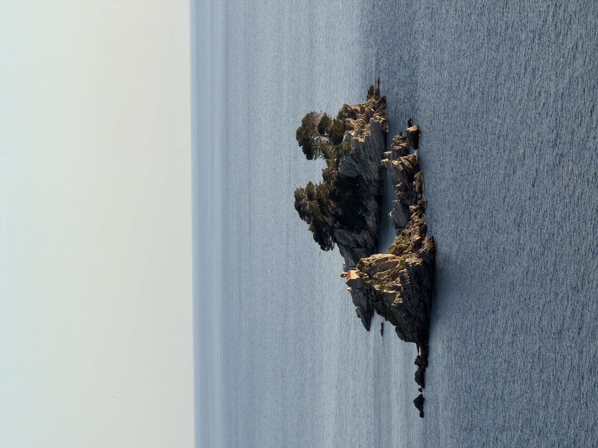 The image captures a rocky islet off the coast of Petrovac centar, Montenegro, surrounded by the Adriatic Sea. The islet features trees and vegetation growing on its surface. A small structure, possibly a chapel or a small building, is visible on the islet. The sea is calm. The sky is a pale, hazy blue.