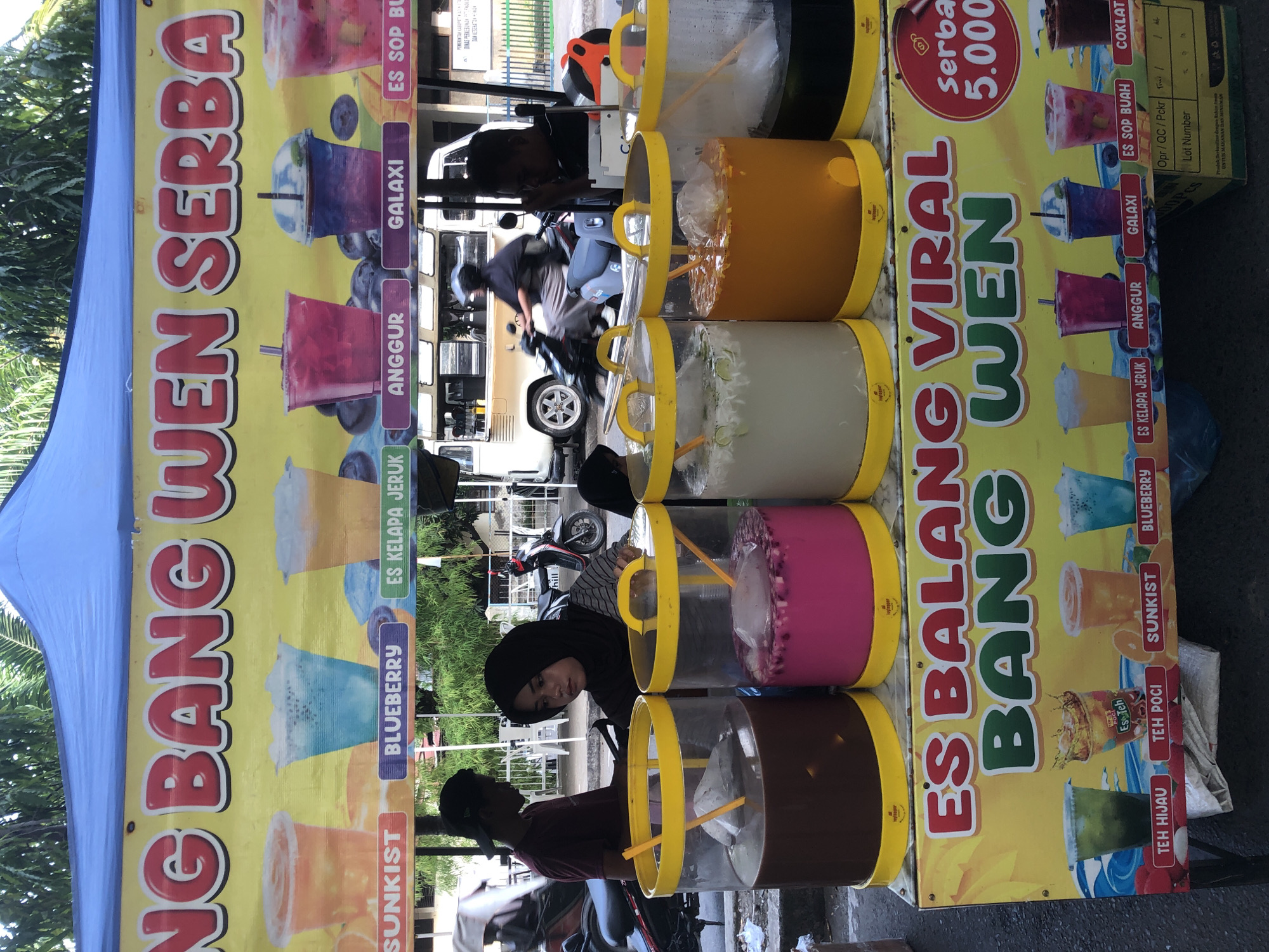 This image captures a street-side beverage stall in Lhokseumawe, Indonesia. The stall is adorned with a yellow banner displaying large, red lettering that reads "BANG WEN SERBA". Below this, a second banner prominently features "ES BALANG VIRAL BANG WEN" with accompanying images of various colorful drinks in cups. A circular red emblem on the right of the banner advertises "Serba 5.000".
In the foreground, large, clear plastic containers filled with different beverages are visible. These containers have yellow lids with handles and yellow spouts at the bottom. The drinks appear to be of various colors, including dark brown, pink, light white, and orange. One container has ice and what looks like fruit pieces inside.
A young woman wearing a black hijab and a striped shirt is leaning over one of the containers, seemingly attending to the drinks. Another person, a male wearing a maroon t-shirt and a black cap, is standing to the left, also near the stall. In the background, a white van with its doors open is parked, and a person on a motorcycle is driving past. The environment suggests a typical street scene in a city, with some greenery and buildings in the distance. The overall impression is of a busy, open-air food and beverage vendor.