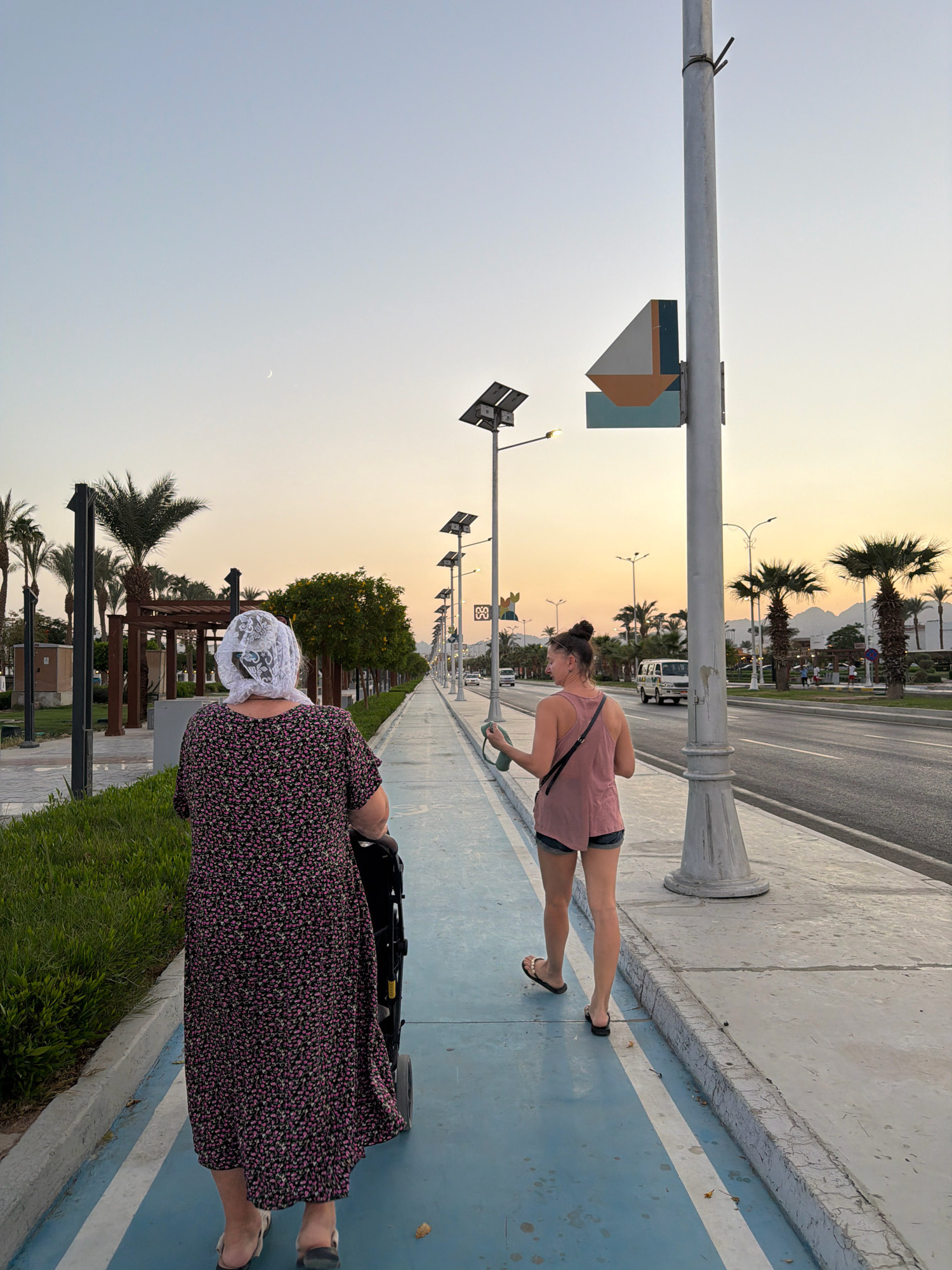 In Sharm El Sheikh, Egypt, a woman wearing a floral dress and a white headscarf walks away from the camera, pushing a stroller along a blue and white striped pedestrian path. To her right, a younger woman wearing a pink tank top and denim shorts walks in the same direction, holding a green bottle and a phone. A tall pole with a decorative sail-like sculpture and a solar panel is positioned between them. The path runs alongside a road with palm trees and buildings in the background, under a pale orange and blue sky with a sliver of moon visible. The sun is setting, casting a warm glow on the scene.