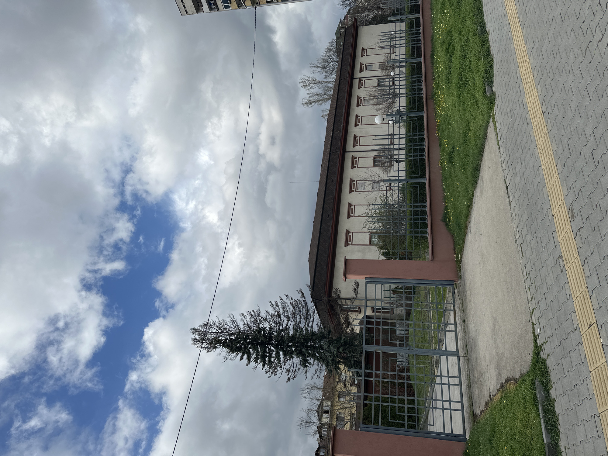 The image depicts a street scene in Sofia, Bulgaria. A long, low-rise building, possibly an institution or apartment block, runs along a street with a grey brick sidewalk marked with a yellow tactile paving strip. The building is light-colored with a dark roof and red window frames. A metal fence borders the building's grounds, with grass and some small trees visible behind it. A tall conifer tree stands near the entrance gate. The sky is partly cloudy with patches of blue. Overhead power lines are also visible. A taller building is partially visible in the background to the top left.