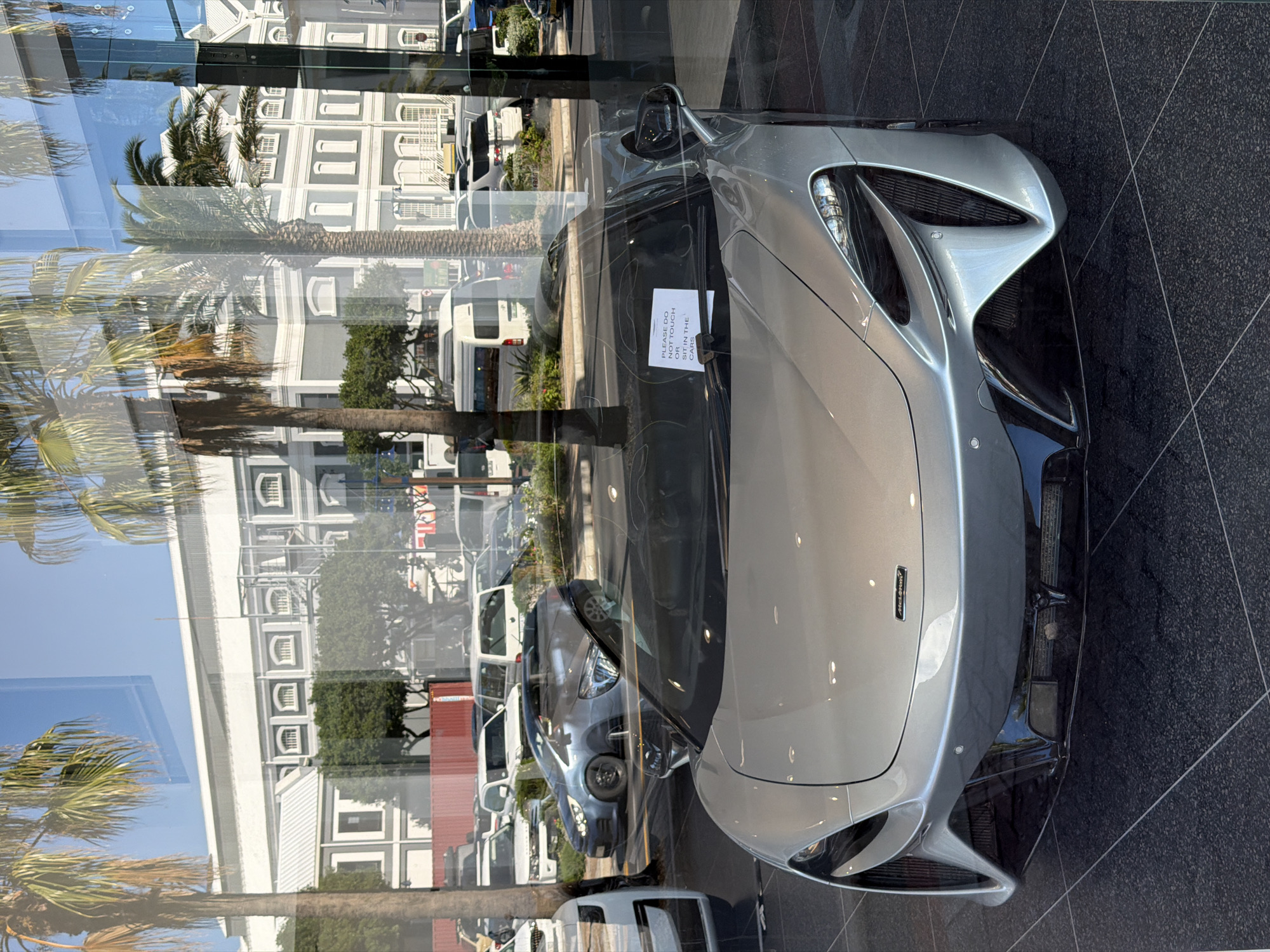 The image shows a silver McLaren sports car parked indoors, viewed through a glass window. Reflected in the window is a street scene with palm trees, a large, white building with multiple windows, and other vehicles parked along the street. A sign is attached to the car's windshield that reads "PLEASE DO NOT TOUCH OR SIT IN THE CARS." The car is parked on a dark grey tiled floor. The reflections make it difficult to pinpoint the exact location, but the architecture and vegetation suggest a location in a coastal urban area, possibly the city of Cape Town, South Africa.