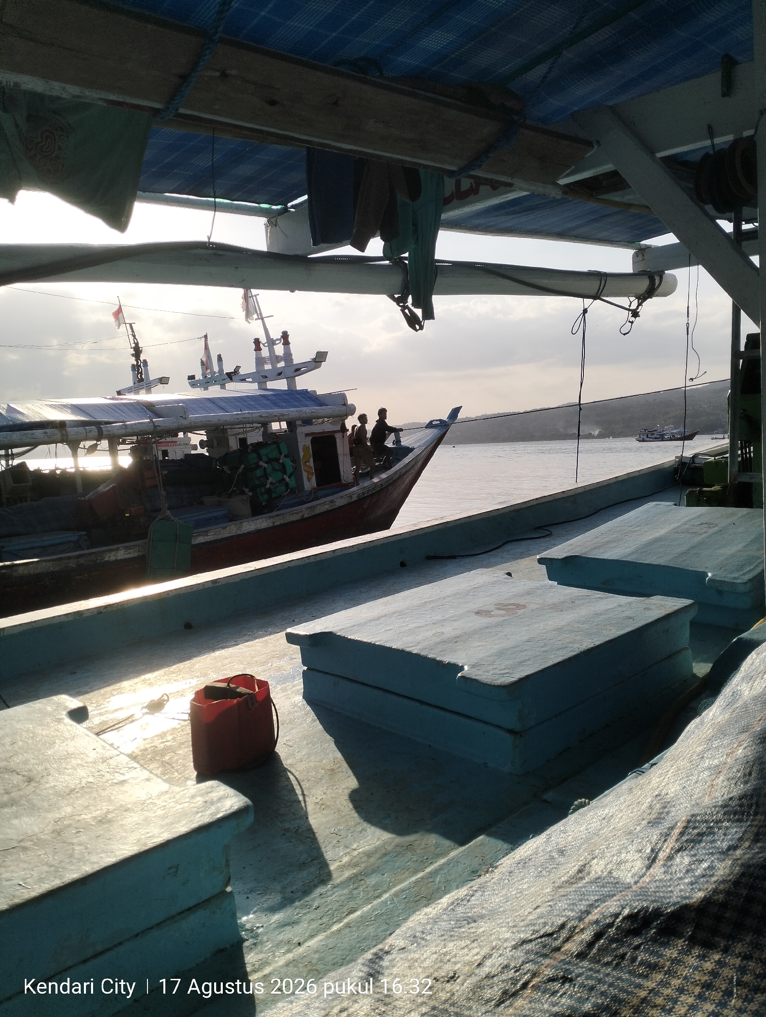 The image depicts a view from the deck of a boat, looking towards another boat docked beside it. The sun is setting or rising, casting a warm glow on the water and the boats. In the foreground, there are several wooden boxes covered in light blue paint, possibly for storage. A red bucket with a dark object inside sits on the deck in the lower left quadrant. The other boat in the background appears to be a fishing vessel with fishing nets and equipment onboard. Several people are visible on the deck of the second boat. The sky is cloudy, with the sun peeking through. The text overlay at the bottom of the image reads "Kendari City | 17 Agustus 2026 pukul 16:32," indicating the location as Kendari City, Indonesia, and the date and time as August 17, 2026, at 4:32 PM. The environment suggests a harbor or coastal area.