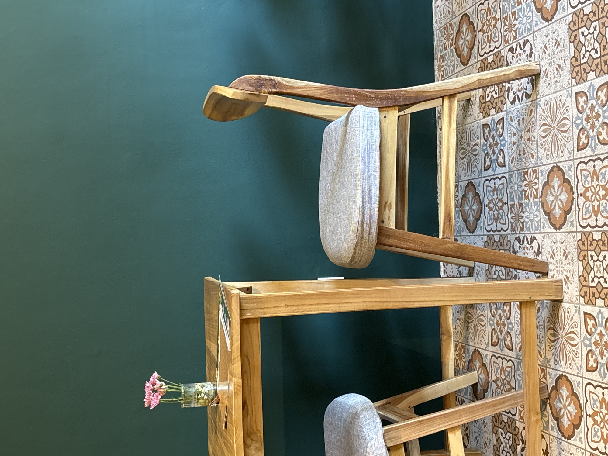 The image features a piece of wooden furniture placed against a backdrop of two different wall surfaces. To the left is a solid green wall, while to the right, the wall is covered in patterned tiles. The furniture appears to be a combination of a rack and chairs, seemingly stacked or intertwined. A small vase with pink flowers is attached to the furniture on the left side, near the green wall. The setting is a room, potentially a home or shop in Kuta Selatan, Indonesia, given the style of tiles often found in Indonesian interiors. There are no people visible in the image.