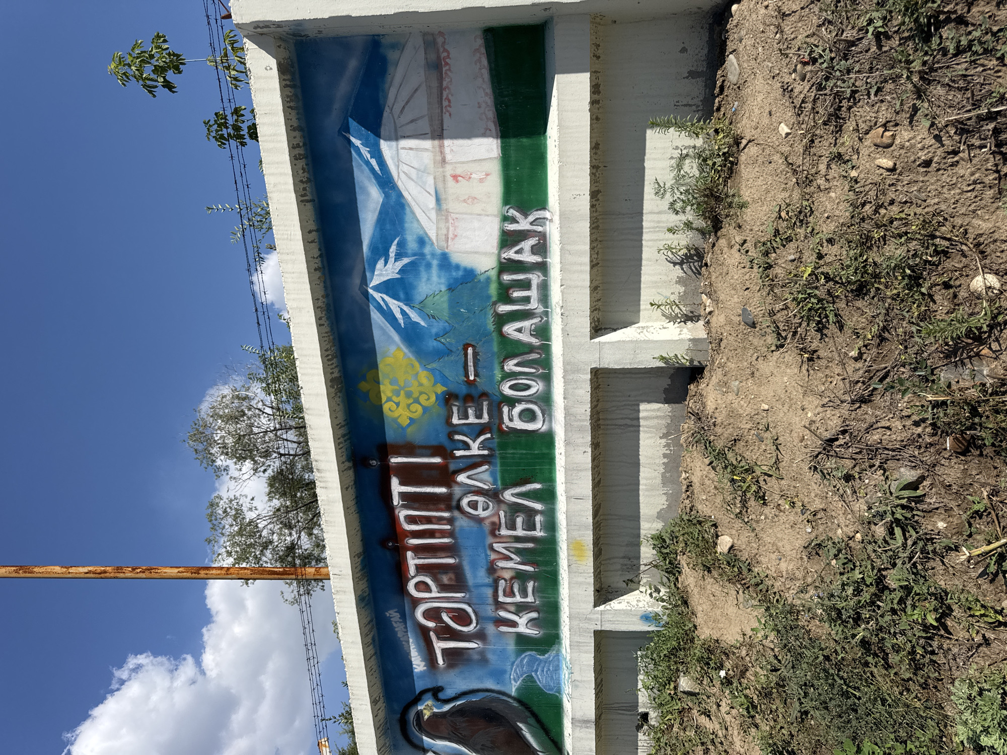 A graffiti-covered concrete wall features a mural depicting a mountainous landscape with a yurt and a bird of prey. The mural includes text in the Kazakh language. The wall is situated in Oskemen, Kazakhstan, as indicated by the graffiti. The scene is set outdoors under a blue sky with scattered clouds. The surrounding area is characterized by dry, overgrown vegetation and dirt. A rusty pole and barbed wire are visible above the wall, suggesting an industrial or utilitarian setting. The graffiti appears to be a public art installation or a form of vandalism.