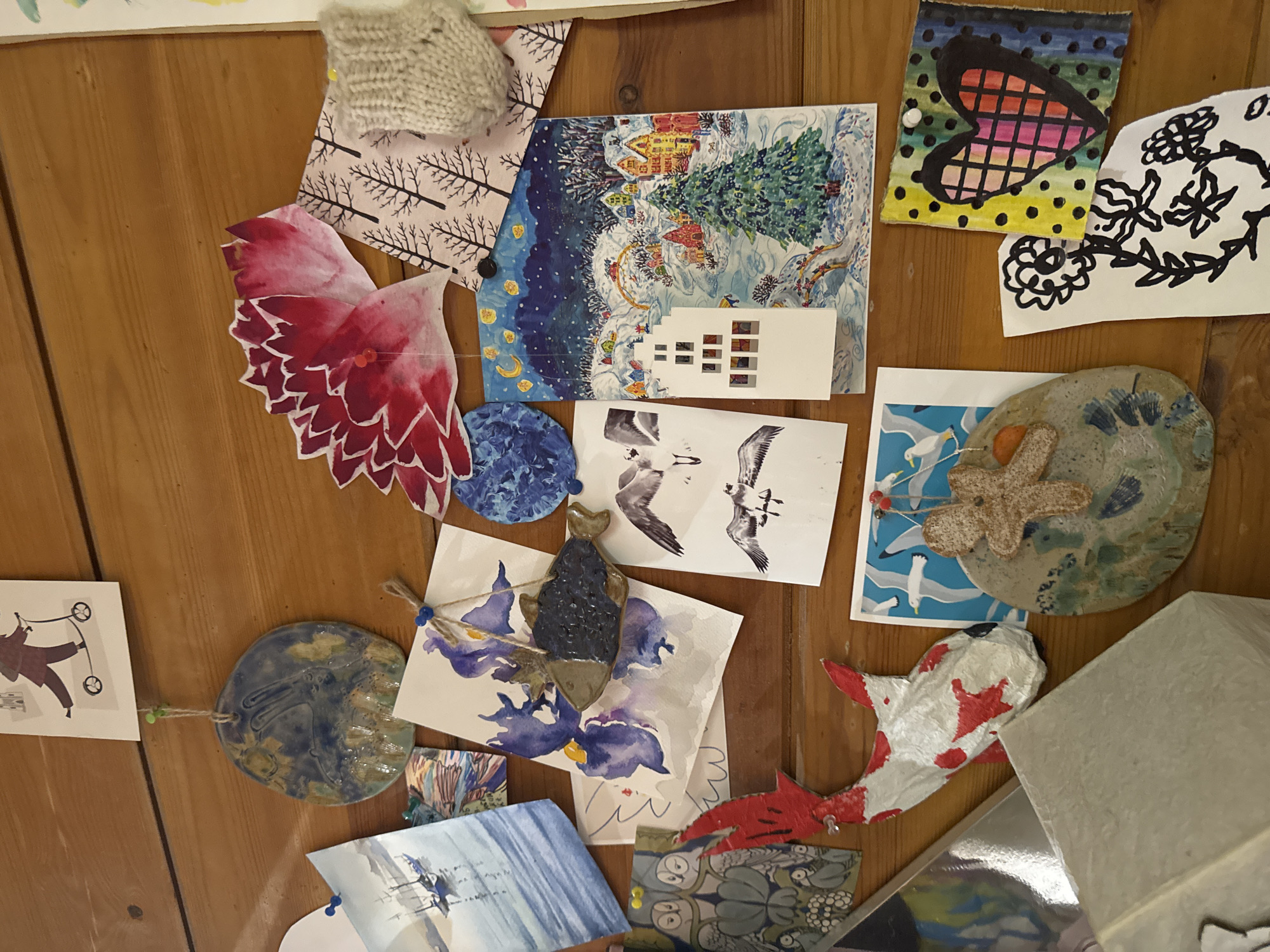 The image displays an overhead view of a wooden surface, likely a table or board, covered with a diverse collection of art, craft items, and postcards, arranged in an overlapping, informal manner. No people are visible.
In the upper left, a light beige knitted swatch is pinned above a piece of paper or fabric patterned with black bare tree branches. Below these, a large, intricately cut-out piece, painted with vibrant red and pink watercolors, possibly resembling wings or a layered flower, is pinned to the surface.
Towards the center top, a rectangular print showcases a whimsical winter village scene with snow-covered houses, green fir trees, and a starry blue night sky. Overlapping this is a white paper cutout depicting the silhouette of multiple interconnected buildings. To the right, a small rectangular drawing on cardboard features a colorful, checkered heart design on a black-dotted background, alongside a white paper cutout with bold black floral patterns.
On the middle right, a circular ceramic piece with a textured, marine-like surface is decorated with a beige starfish and a small red bead. Below it lies a postcard or print depicting white seagulls flying over blue water. In the bottom right, a sculpted fish, possibly papier-mâché, painted white with red spots resembling a koi carp, is visible near a light gray box.
The bottom center and left areas feature a small, dark blue ceramic fish charm hanging by twine, a watercolor painting of large purple irises, and another small, oval ceramic piece with blue-green glaze and an embossed design, also hanging by twine with green beads. A small watercolor painting in blue tones and a card featuring a stylized figure pushing a stroller complete the collection.
The scene suggests an artistic or personal display. No specific activity is occurring, and there are no direct visual cues for time of day or external weather. No discernible text is clearly visible on any of the items. While the context of Moscow, Russia, is provided, the image itself does not contain specific visual identifiers for this location.
