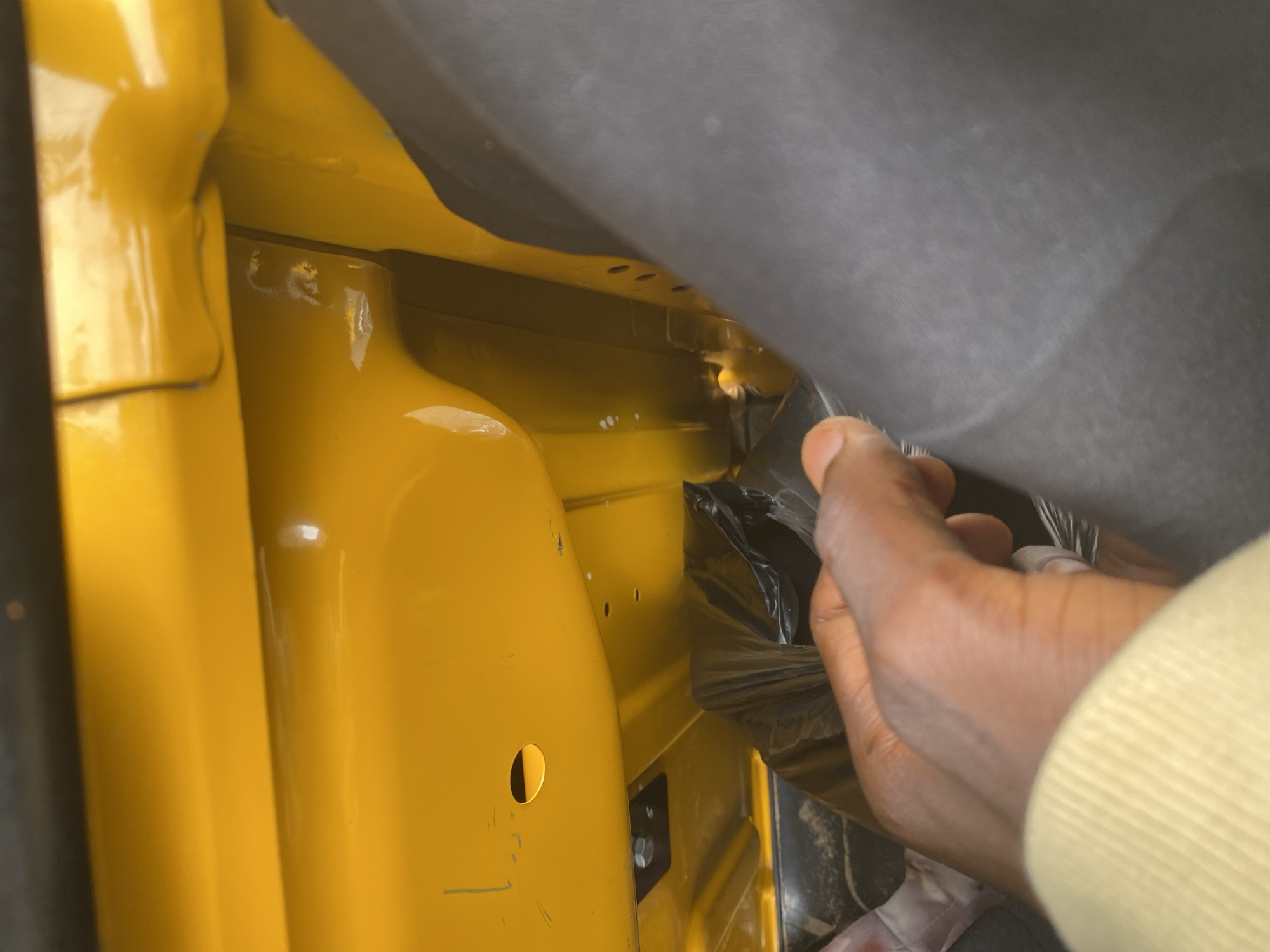 The image shows a close-up view of a person's hand reaching into a yellow-painted compartment, possibly within a vehicle. The hand, which appears to be of dark skin tone, is holding and manipulating a dark, opaque plastic material. The compartment itself is made of painted metal, with some visible bolts and circular openings. A gray fabric or material is also present, obscuring part of the background. The lighting suggests an indoor or shaded environment, with some reflections on the yellow surface. The overall impression is one of someone working on or inspecting the interior of a mechanical or vehicular component. The context provided indicates the location is Jalingo, Nigeria. No visible text is present in the image.