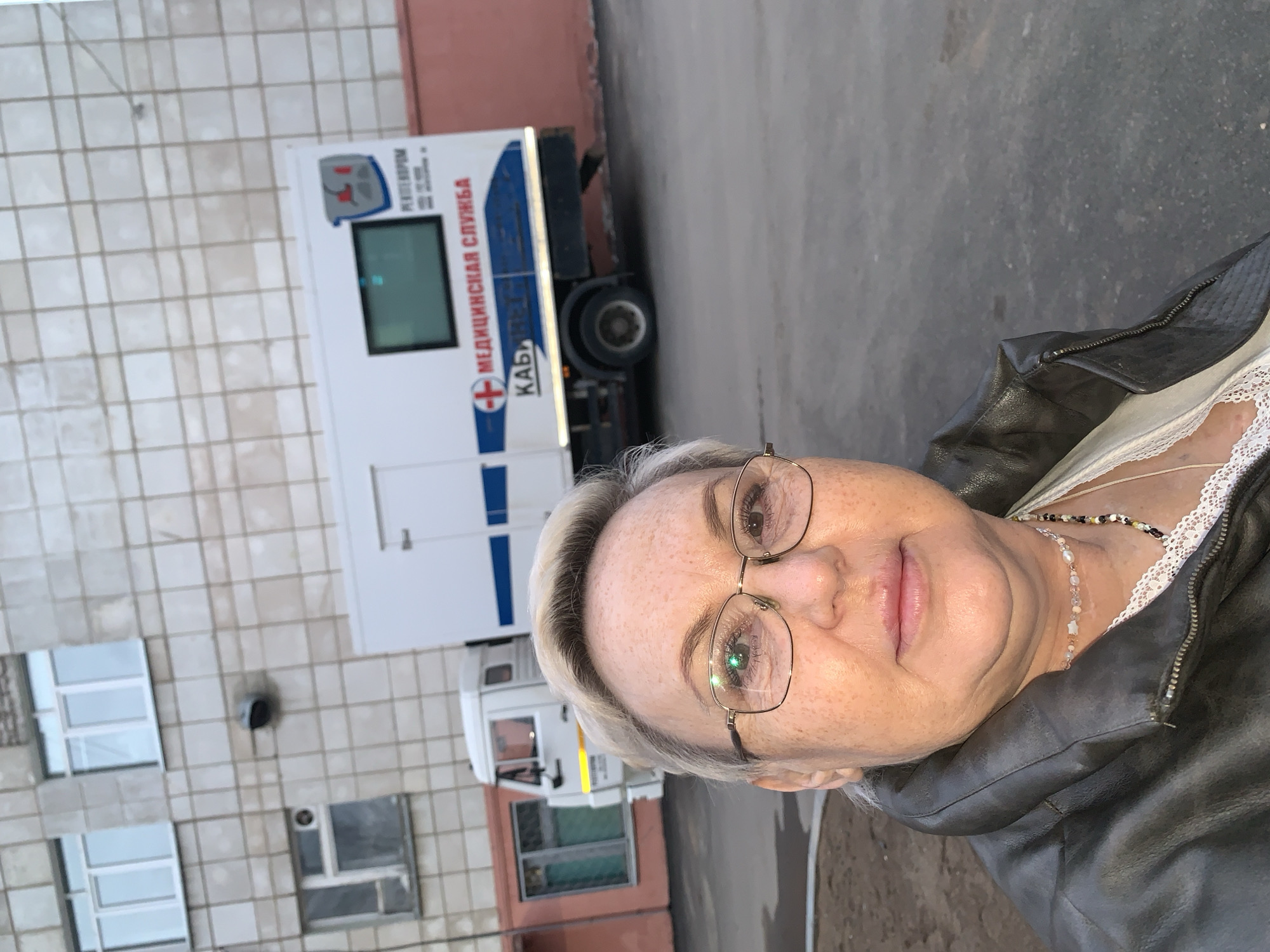 A woman with short gray hair and glasses stands in front of a white truck labeled "Медицинская Служба (Medical Service) КАБ" in the city of Chaikovsky, Russia. She wears a dark jacket over a white top and a necklace. The truck is parked near a building with multiple windows and a pink wall. The environment is urban, with pavement visible.