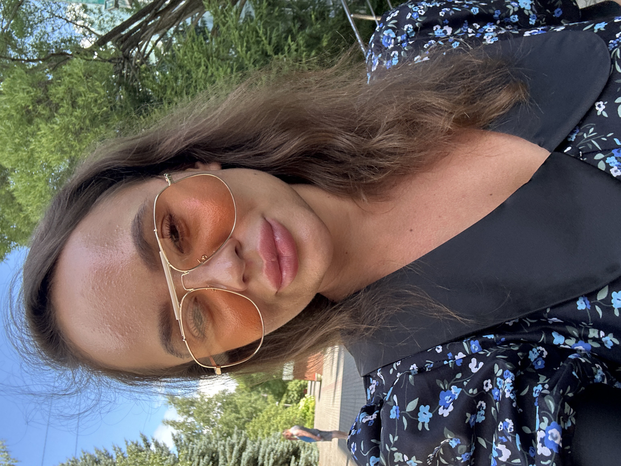A woman wearing sunglasses and a floral dress is posing for a selfie outdoors. She has long, wavy brown hair and is smiling. In the background, there are trees and a man walking away from the camera. The location is the city of Duvankul', Russia.