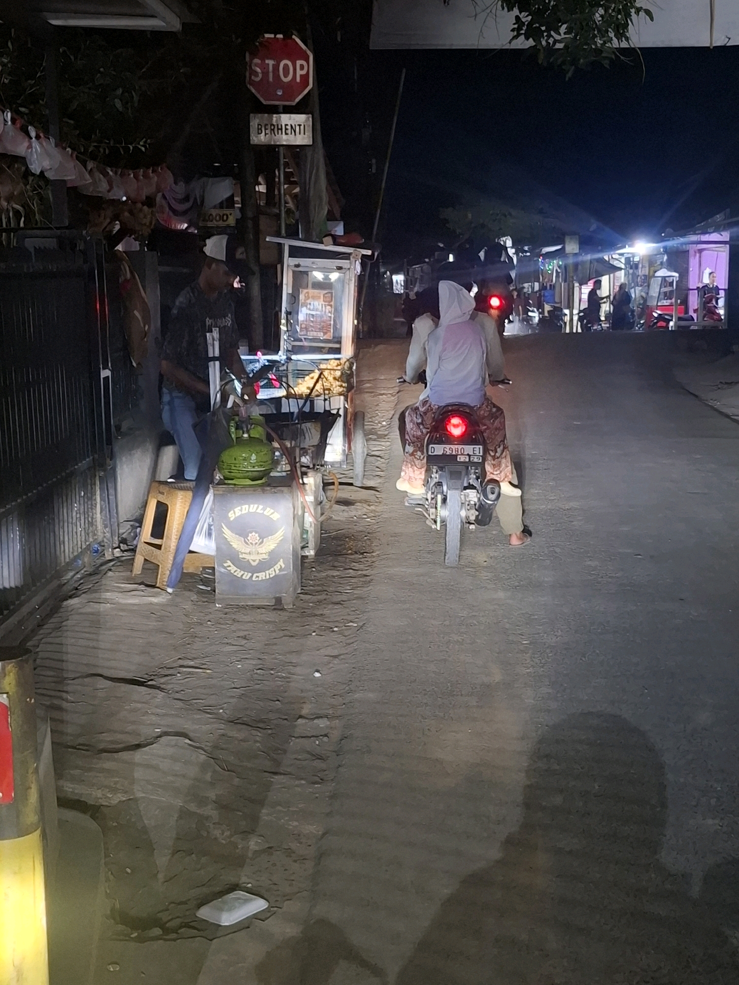 This is a night-time, street-level photograph taken in Karangtengah, Indonesia. The image depicts a street scene with a food cart on the left, displaying a sign with text that includes "SEDULUR TAHOE CRISPY". A person wearing a white hood and patterned pants is riding a motorcycle in the center of the frame, moving away from the camera. The motorcycle has a license plate with the characters "D 6980 EI". To the left of the food cart, a person wearing a dark shirt and a white cap stands near the cart. In the background, several other food stalls and buildings are visible, illuminated by artificial lights, suggesting a commercial or bustling area. A "STOP" sign with "BERHENTI" below it is also visible on a pole near the food cart. The road surface appears to be asphalt, and shadows are cast on the ground, indicating the time of day and artificial lighting.