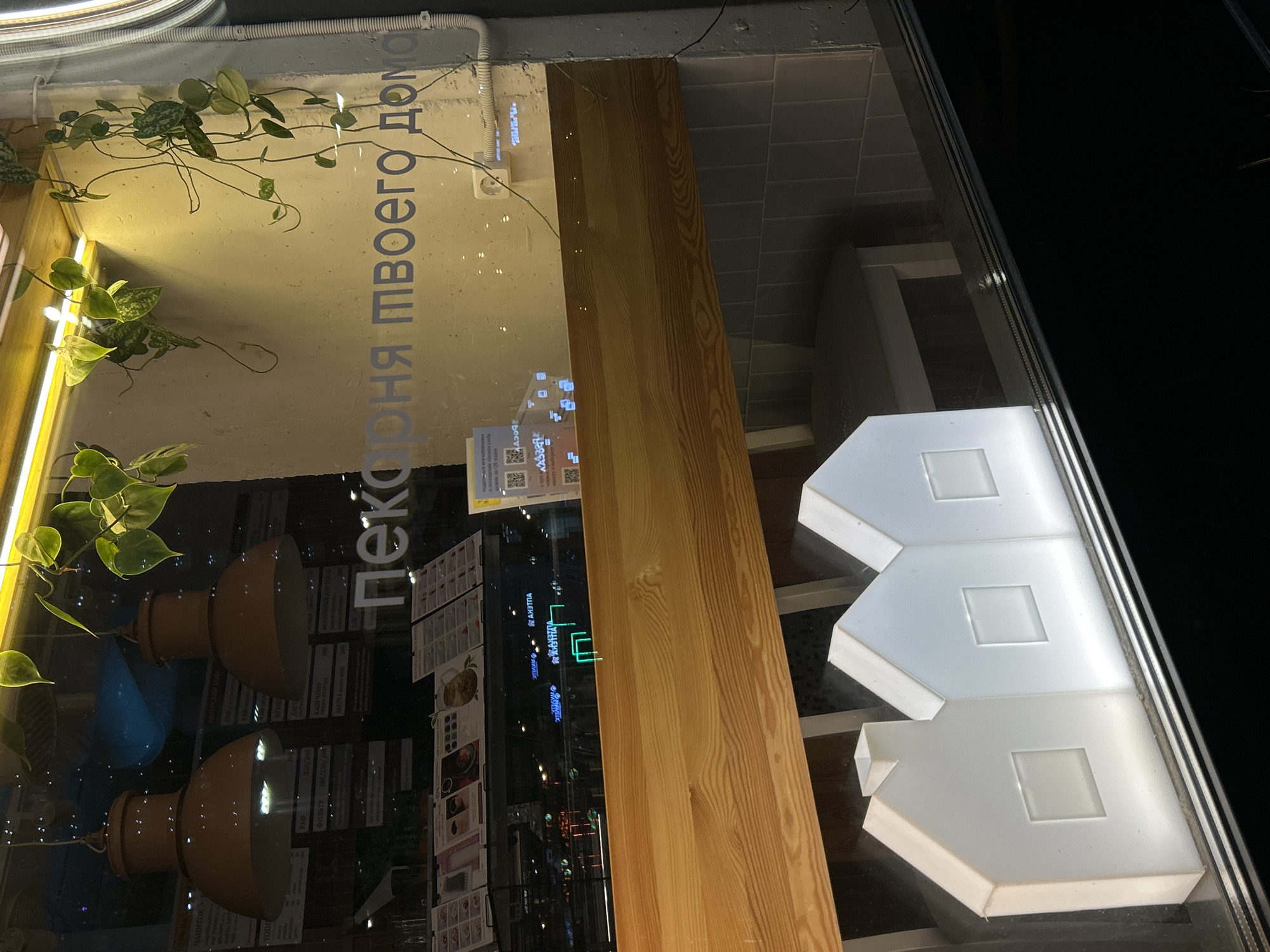 This media file captures a nighttime view looking through the window of a shop, likely a bakery, in Saint Petersburg, Russia. The scene is static, devoid of any human activity.
Inside the well-lit establishment, several elements are visible through the glass: green plants with trailing vines hang along the upper left, illuminated by a linear light source beneath them. Further inside, two brown, bowl-shaped pendant lights are suspended. A light-colored wooden panel or pillar runs vertically through the middle of the frame, and to the far right, three white, stylized house-shaped light fixtures with square cutouts are arranged.
Prominently displayed on the glass, etched or decaled, is the Russian text "Пекарня твоего дома," which translates to "Your Home Bakery." The glass also bears numerous reflections. These include rectangular menu-like items with discernible text such as "HAWATOK," "RED BIRD," "ШАУРМА" (Shawarma), "ФАЛФЕЛЬ" (Falafel), and various price indications. Multiple QR codes are also reflected. Additionally, the word "АПТЕКА" (Apteka), meaning "Pharmacy," is reflected several times, suggesting a nearby pharmacy sign is bouncing off the window.
The dark background outside, visible in reflections, confirms the time of day as night, highlighting the warmth and light of the interior.