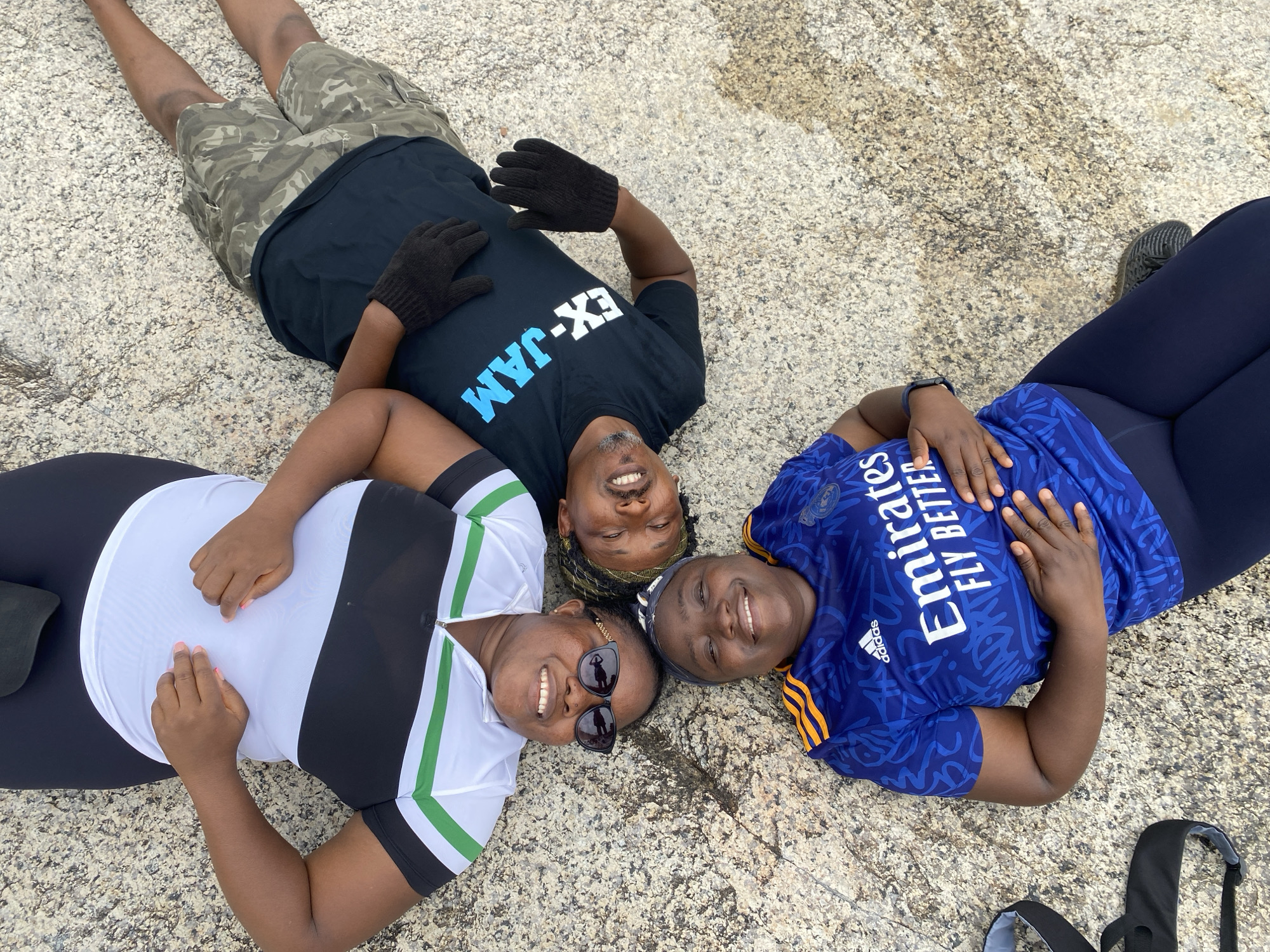 Three adults are lying on a textured stone surface outdoors, arranged in a roughly triangular formation. The person at the top wears a black t-shirt with "EX-JAM" printed on it, camouflage shorts, and gloves. The person on the left wears a white shirt with black sides and green stripes. The person on the right wears a blue soccer jersey with "Emirates FLY BETTER" printed on it and dark leggings. All three are smiling and appear relaxed. The location is Ganawuri, Nigeria.