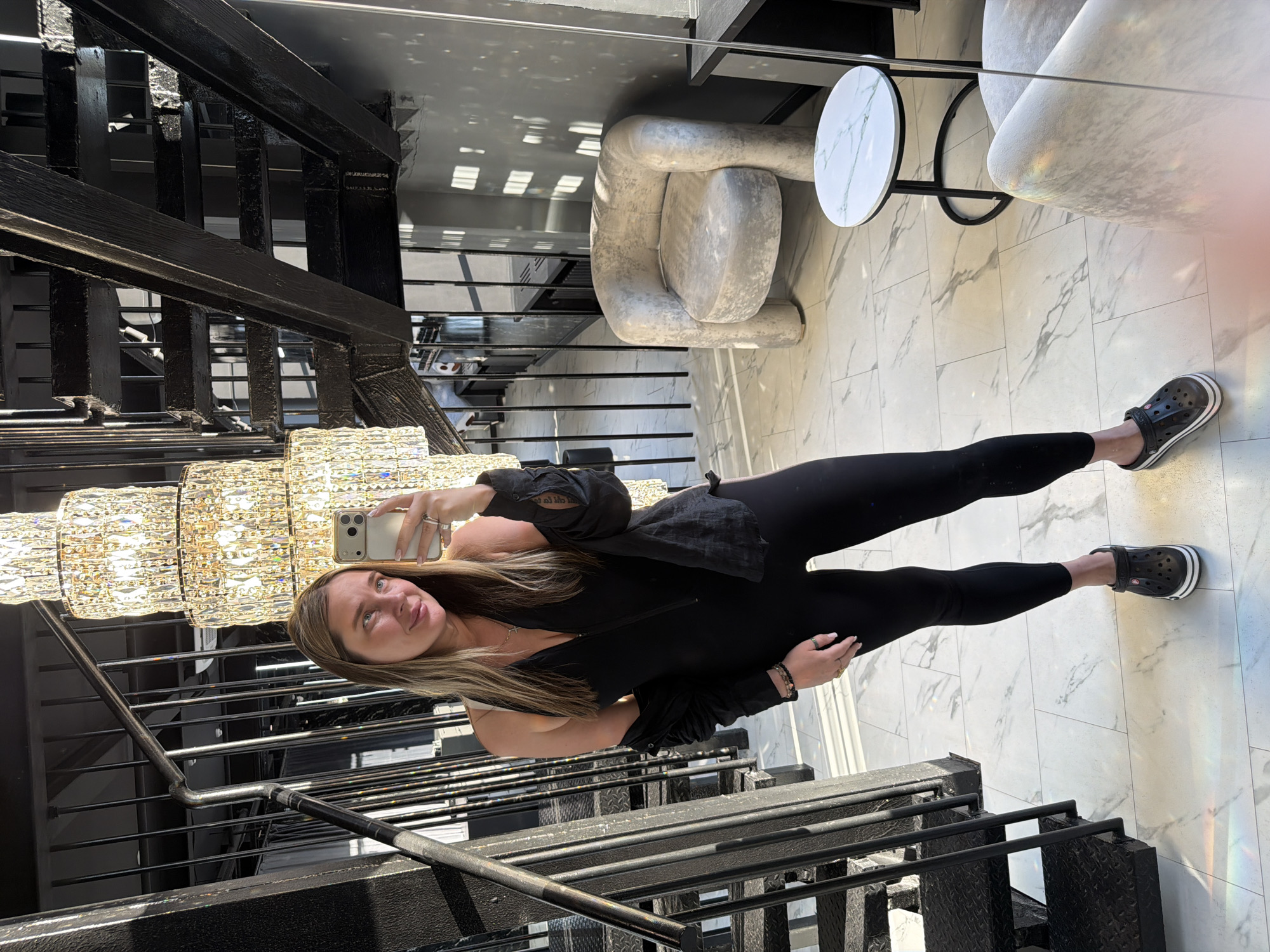 A fair-skinned woman is taking a selfie in front of a large crystal chandelier and black metal staircase. She is wearing black leggings, a white tank top, a black zipper jacket, and black crocs. She is standing on a white marble floor. In the background, there is a white textured chair and a small round white table. The location is not readily identifiable as Moscow, Russia from the visual information provided.