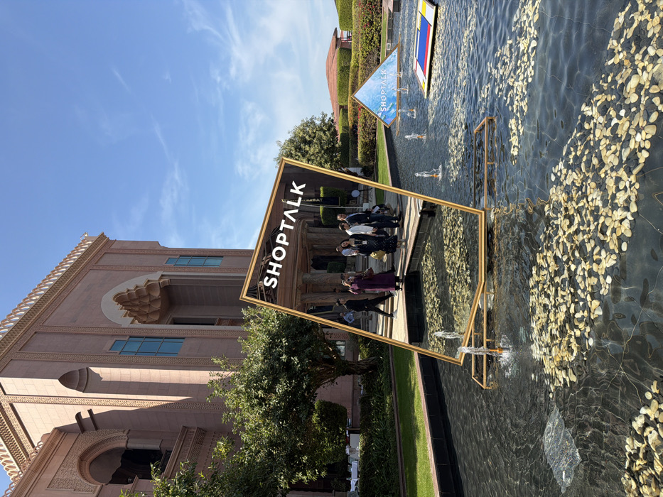 The image depicts an outdoor scene at the Emirates Palace Mandarin Oriental in Abu Dhabi, United Arab Emirates. In the foreground, a large, rectangular, gold-framed sign or mirror titled "SHOP TALK" in white capital letters is positioned at an angle, partially submerged in a dark-bottomed fountain. This reflective surface displays the palace entrance area and several individuals. The fountain contains numerous scattered light-colored pebbles and features multiple active water jets. Further to the right within the fountain, a blue and white pyramid-shaped sign also displays "SHOP TALK," alongside a floating rectangular object patterned with primary colors and black lines, resembling a Piet Mondrian artwork.
The background is dominated by the Emirates Palace, a grand, sand-colored building exhibiting intricate Islamic architectural elements including ornate archways, window designs, and a visible upper floor structure. Manicured green trees and hedges are situated between the fountain and the palace facade. The sky above is clear blue with sparse white clouds. The individuals visible near the palace entrance and reflected in the sign are dressed in various business casual to semi-formal attire.