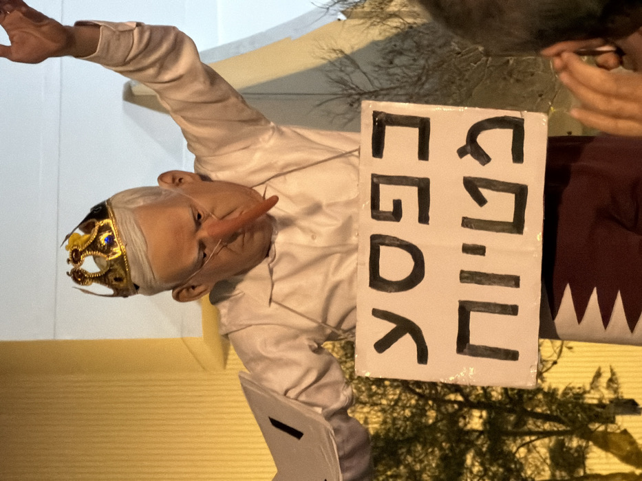 The image displays a person dressed in a costume, participating in what appears to be a protest or satirical demonstration. The individual is depicted as a caricature, wearing a gold crown adorned with blue gems, and a prominent, elongated, light orange/reddish pointed nose, reminiscent of Pinocchio. They also wear a white head covering or mask partially obscuring their lower face, and glasses. Their hair appears light-colored, possibly white or grey.
The person is wearing a long-sleeved, white collared shirt. Their left arm is raised high and slightly bent, with the hand open. In their right hand, they hold a rectangular white sign with black text. The text is in Hebrew and reads: "שקרן" (Shekeran), meaning "Liar," followed by "לכלא" (Lechela), meaning "To jail" or "Jail him." Therefore, the sign's message is "Liar, to jail."
In the background, on the right side of the image, a dark, possibly maroon or dark red fabric with distinctive white triangular or jagged patterns along its edge is visible. This pattern strongly resembles the design elements of the Qatari flag. Above this, and behind the sign, are some dark, leafy branches, suggesting the presence of trees or bushes, possibly indicating an outdoor setting. On the left side of the image, a plain, light-colored wall or surface is visible, which could be white or a light yellow.
The lighting has a warm, yellowish hue, suggesting it might be nighttime with artificial illumination, or an indoor setting with specific lighting. The overall impression is one of political commentary or protest, using satirical elements (Pinocchio nose, crown) to convey a message about dishonesty and accountability.