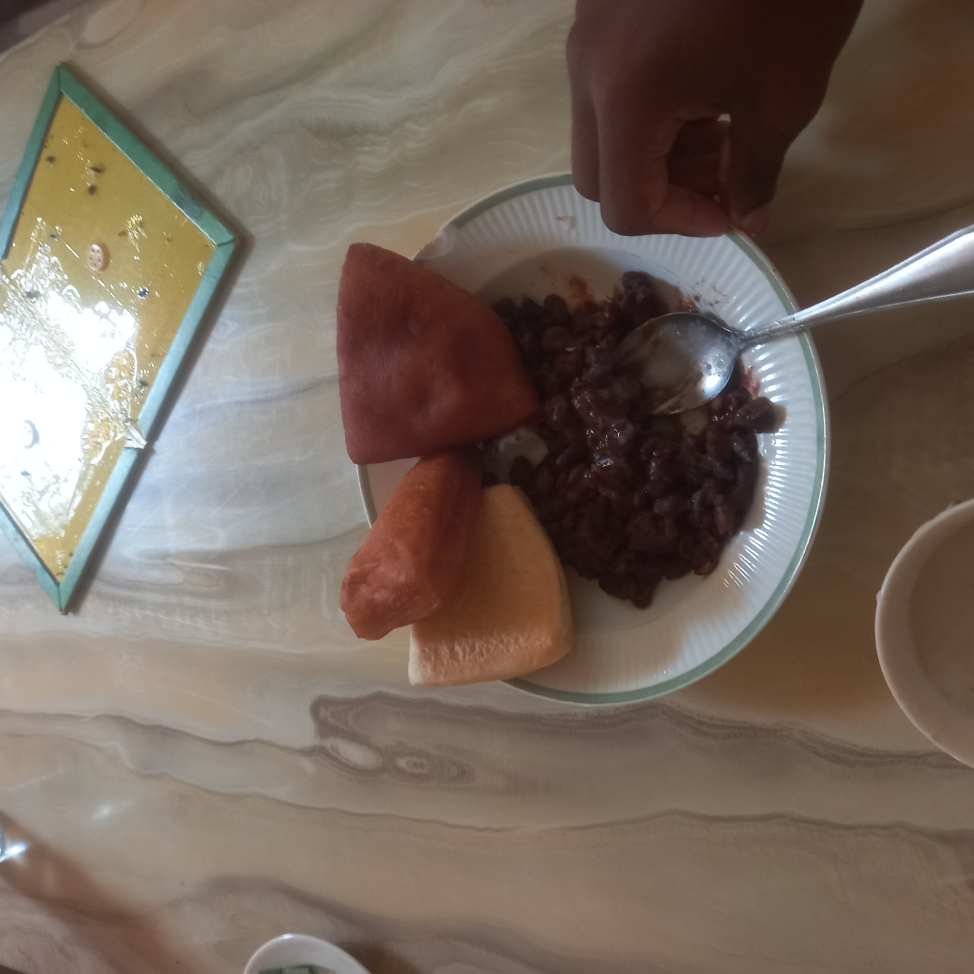 The image depicts a close-up view of a meal served on a white plate with a green rim, placed on a marble-patterned table. The meal consists of dark beans, possibly kidney beans or black beans, swimming in a dark sauce. Alongside the beans are three pieces of fried dough, two of which are triangular and reddish-brown, while one is a lighter, golden-brown. A spoon rests on the plate, partially submerged in the beans, with a hand reaching from the right side of the frame to hold the edge of the plate. In the upper right portion of the image, a yellow object with a green frame is visible, appearing to be a fly trap or a similar sticky surface, with several dark specks on it. The background is dominated by the marble-patterned table, with hints of a light-colored tablecloth underneath in some areas. The lighting suggests an indoor setting, possibly a restaurant or home in Zanzibar, Tanzania.