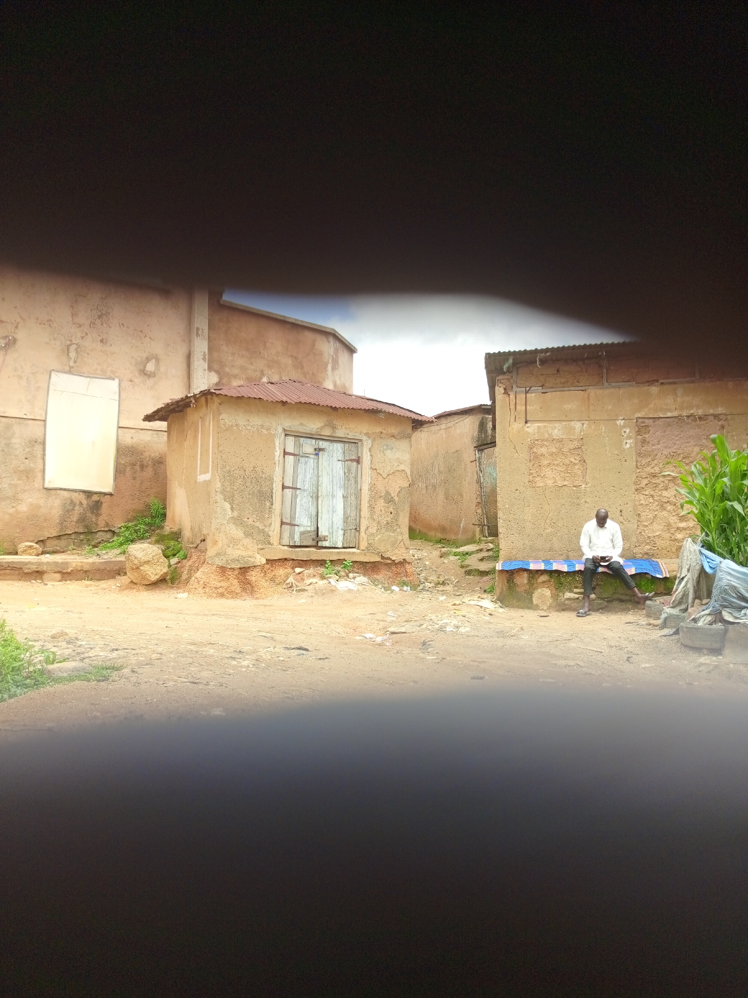 In Jos, Nigeria, a man in a white shirt and dark trousers sits on a blue and white patterned mat laid over a low concrete structure. He is looking down at a black object, likely a phone, in his hands. He is positioned in front of a weathered, sand-colored building with a corrugated metal roof. To the left of the building is a smaller, similar structure with wooden double doors. The ground is unpaved, appearing to be dirt and sand, with some scattered debris. Green vegetation, including what appears to be corn stalks, is visible to the right of the man. The overall environment suggests a rural or semi-urban setting with simple structures and a dusty, natural ground cover. The image is partially obscured by dark, blurred areas at the top and bottom, potentially from a lens or frame.