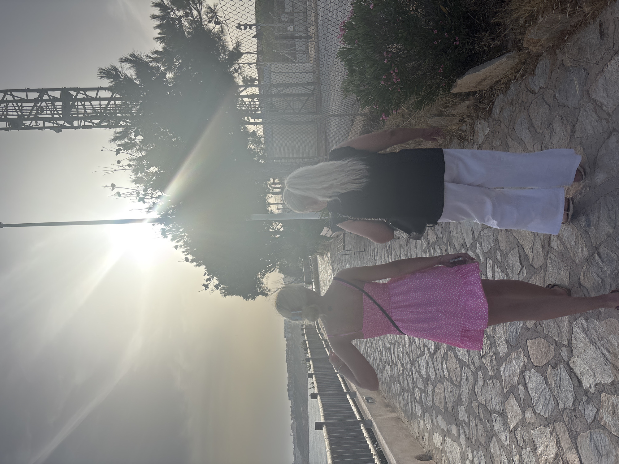 Two individuals are walking away from the camera on a cobblestone path next to a body of water. The person in the foreground, on the left, is wearing a pink, polka-dot sundress and has blonde hair pulled back. The person behind them, on the right, is wearing a black top and white wide-leg pants, with long blonde hair in a ponytail. A wooden fence runs alongside the path, bordering the water. In the background, there is a cell tower and some vegetation. The sun is low in the sky, casting a bright glare and creating lens flares. The overall setting appears to be a coastal area. This scene is likely located along the coast of Orihuela, Spain, potentially in a town like Torrevieja, which is part of the Orihuela municipality and known for its coastal promenades.