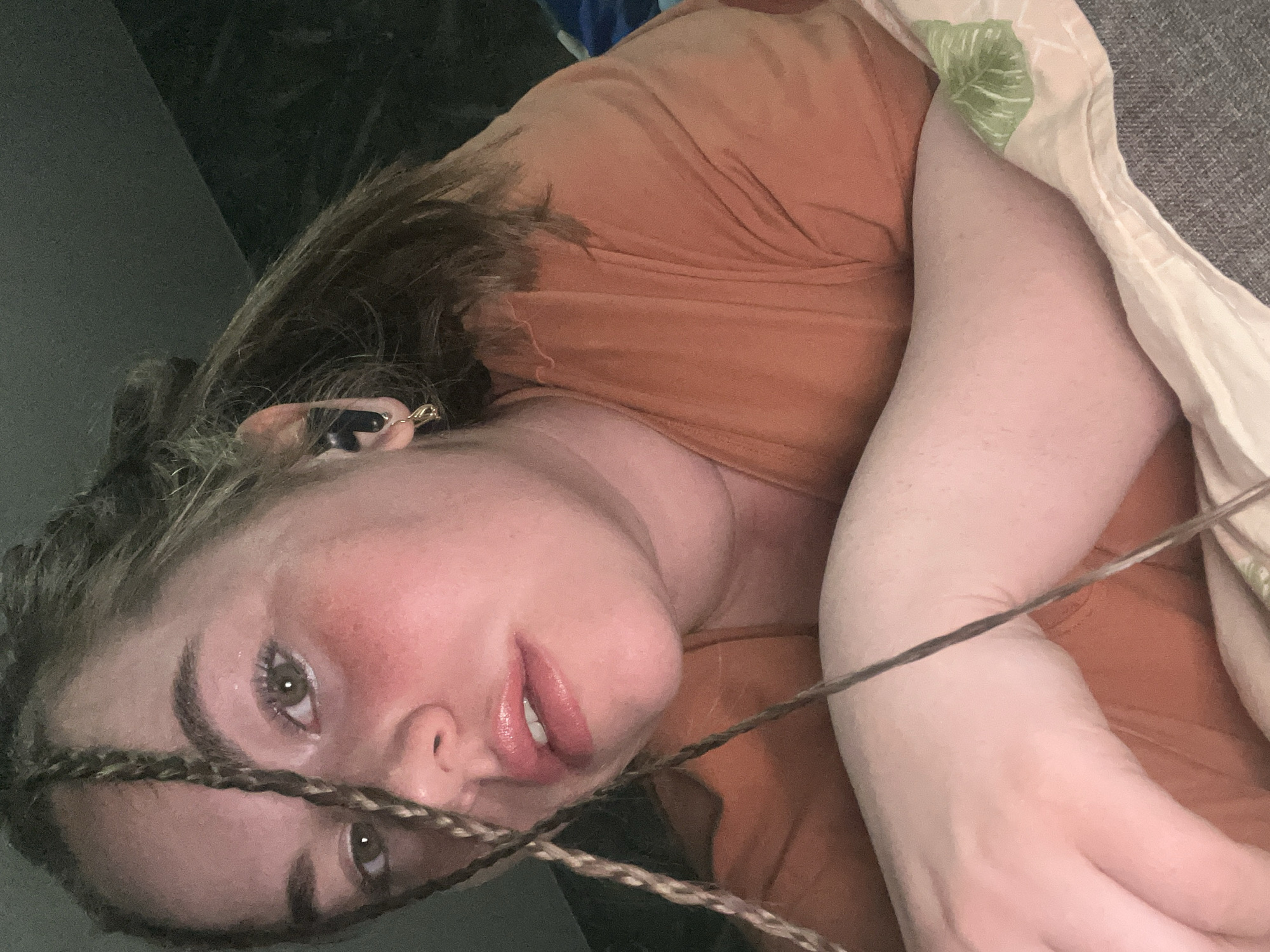 The image depicts a close-up, angled view of an individual, presumably female, lying prone and facing the camera. The subject has light brown hair, some sections of which are styled into thin braids, with one braid falling across the left side of the face. Visible facial features include green eyes, a reddish complexion on the cheeks, and slightly parted lips with a glossy finish, revealing some upper teeth. A black in-ear earbud is present in the subject's right ear, accompanied by a small gold-toned hoop earring. The individual is wearing an orange or rust-colored v-neck t-shirt. The forearms are visible in the lower foreground, resting on a surface. The immediate environment beneath the subject appears to be a light-colored patterned fabric, possibly a sheet with a green leaf motif, which in turn rests on a textured grey upholstered surface, likely a bed or sofa. The background is dark and indistinct, suggesting an indoor setting with low ambient light, and direct illumination on the subject's face. This scene is located in Chaikovsky, Russia.