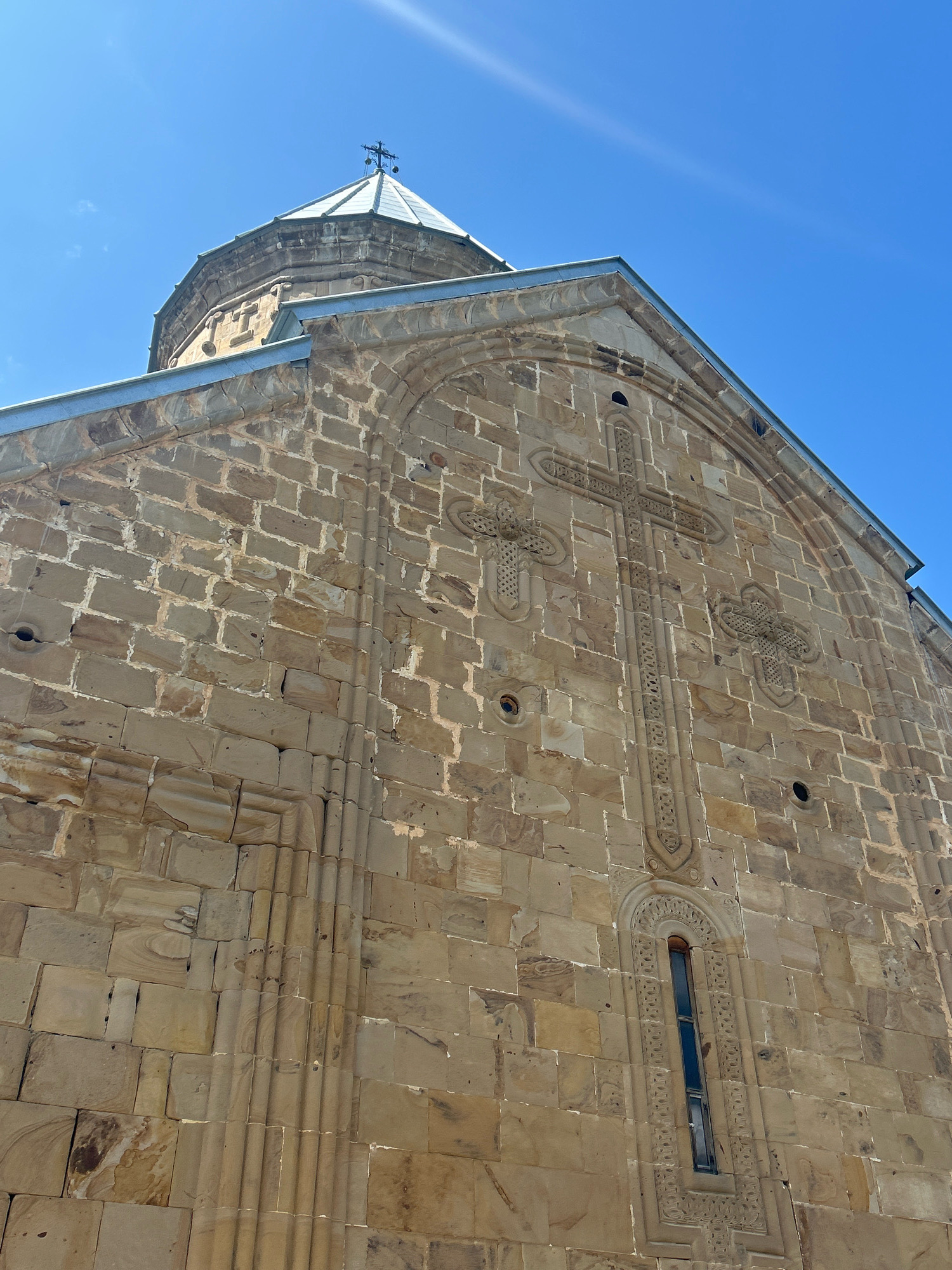 The image shows the exterior of a stone church in Shalikiantkari, Georgia, under a clear blue sky. The architecture is characterized by rough-hewn stone blocks and intricate carvings. Prominently featured are large crosses etched into the facade, one central and vertical, with smaller ones to its sides. A narrow, arched window with ornate detailing is visible below the central cross. The church has a conical dome topped with a cross. The stone is weathered and varies in color from light tan to brown, with visible mortar between the blocks. The perspective is upward-looking, emphasizing the height and grandeur of the building.