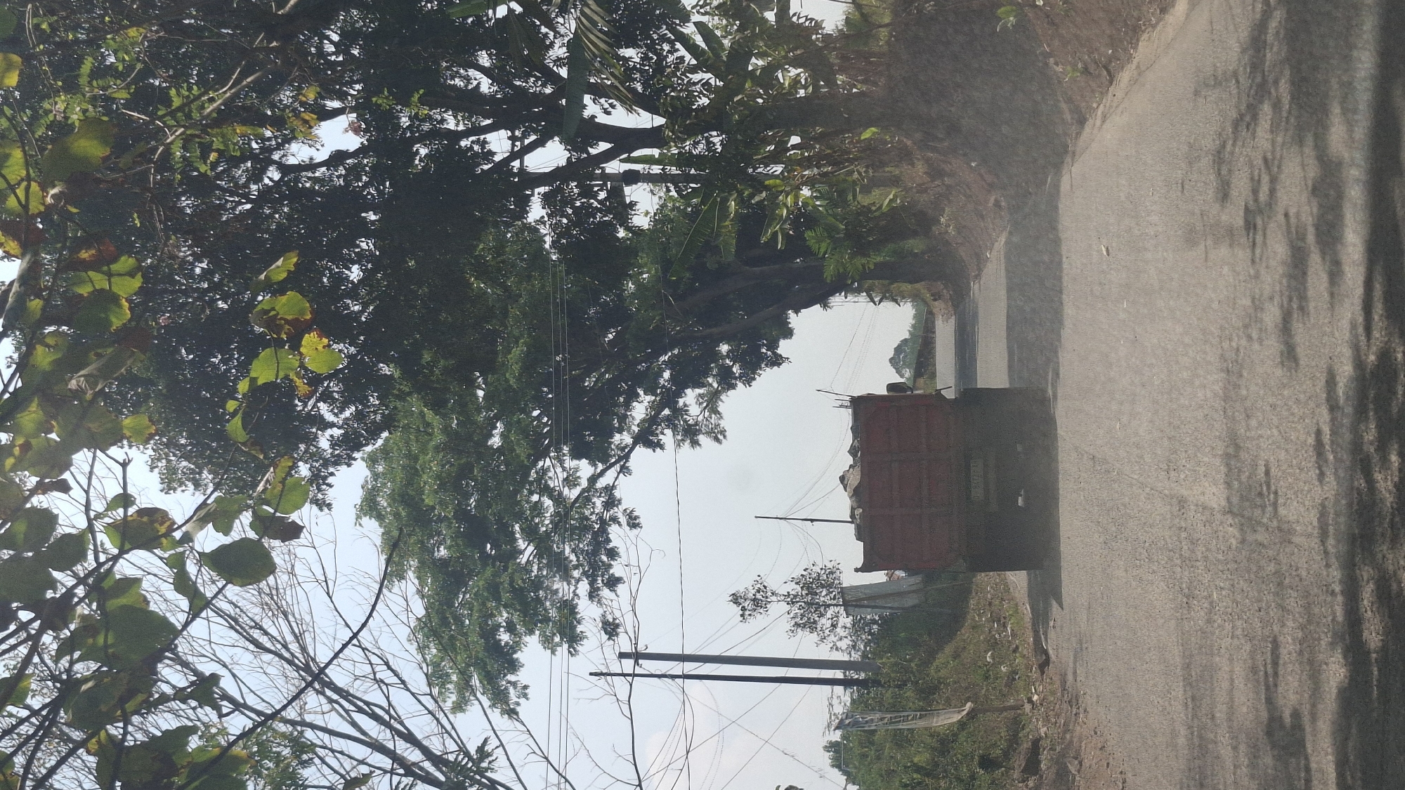 A red truck with a full load of debris is driving down a paved road in Cibinong Hilir, Indonesia. The truck is in the center of the frame, moving away from the viewer. The road is lined with trees and vegetation, with power lines visible in the background. The sky is overcast. The image appears to have been taken from a moving vehicle, as indicated by the motion blur and the angle of the shot.