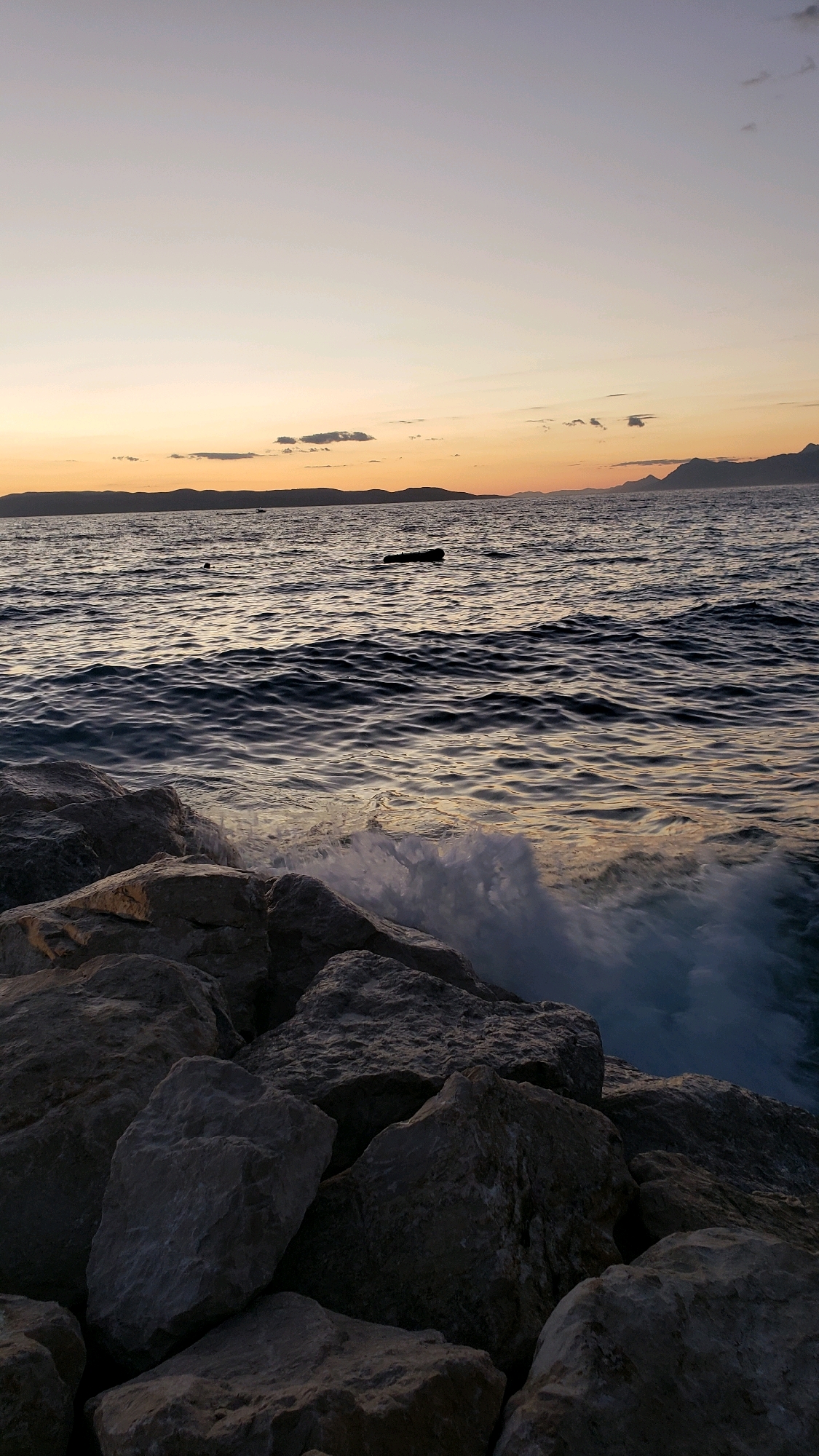 This is a vertical, eye-level shot of a rocky shore at sunset. The foreground is dominated by large, grey rocks, some of which are wet from the sea. The sea itself is dark blue with visible ripples and waves, with white foam splashing against the rocks on the right side of the frame. In the middle ground, there are a few dark objects floating on the water, one of which resembles a small boat or buoy. The background features a distant coastline with rolling hills and mountains, bathed in the warm hues of the setting sun. The sky transitions from a pale orange and yellow near the horizon to a soft grey at the top of the frame. The overall impression is one of a serene, natural landscape at dusk. This scene is likely located along the coast of Općina Tučepi, Croatia, a region known for its beautiful Adriatic coastline and mountainous backdrop.