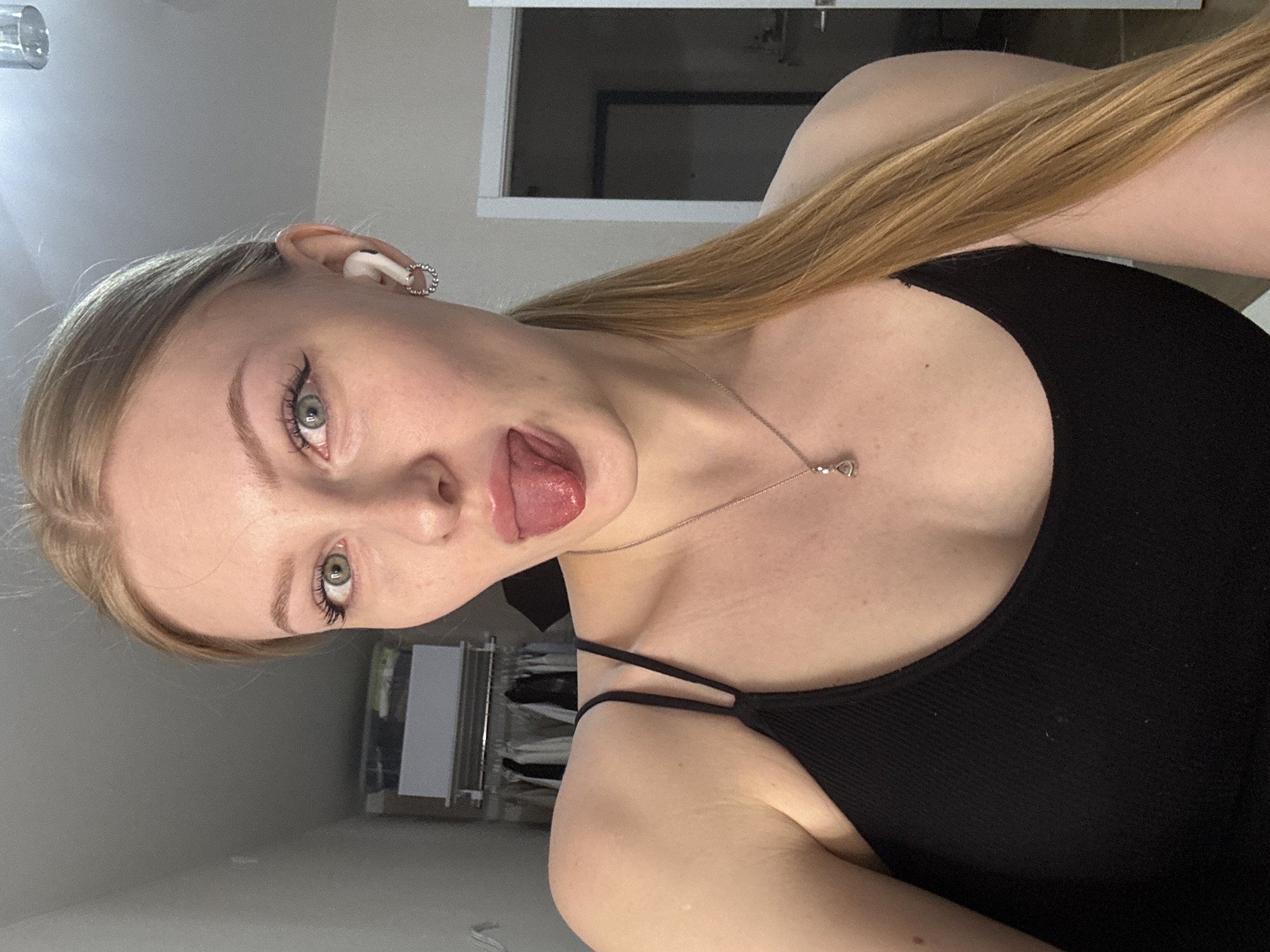 A woman with blonde hair is wearing a black tank top. She is sticking her tongue out and has an earring on her ear. She also has a necklace on. In the background, there is a closet and a window. The location is not evident from the image.