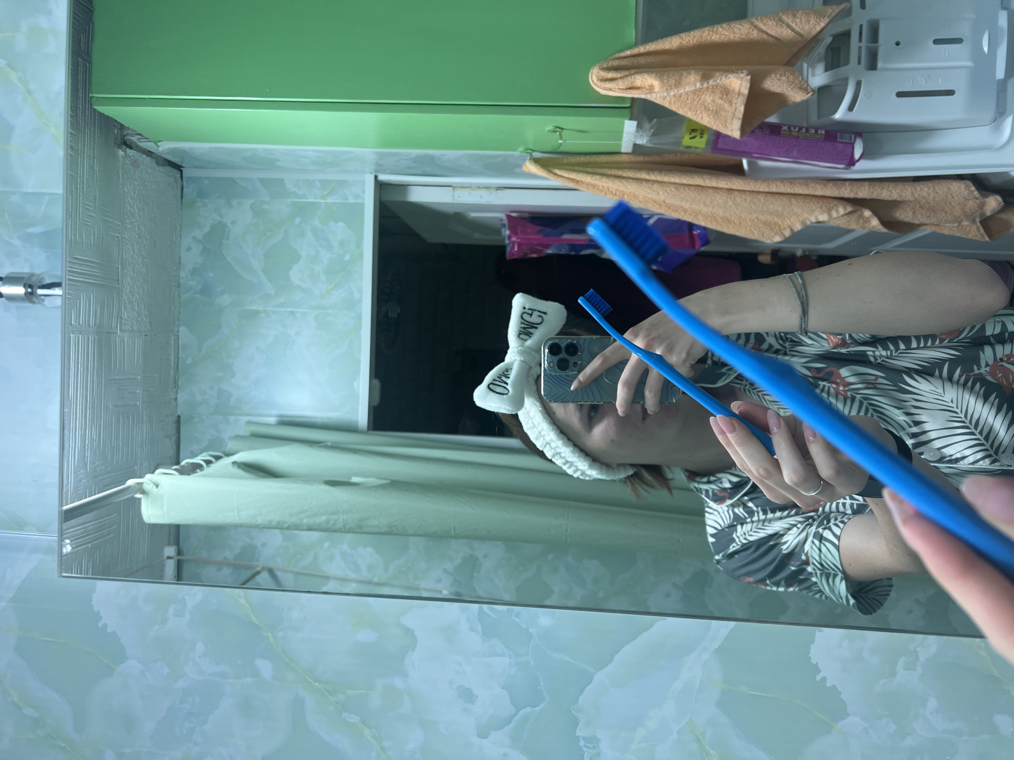 A person is taking a mirror selfie in a bathroom. They are wearing a white headband with a bow and a tropical print pajama top. They are holding two blue toothbrushes and a smartphone. The bathroom features mint green tiled walls, a green cabinet, and several towels hanging on the wall. The scene is consistent with a residential bathroom. The provided crops do not offer enough information to determine the exact location within the city of Chaikovsky, Russia.