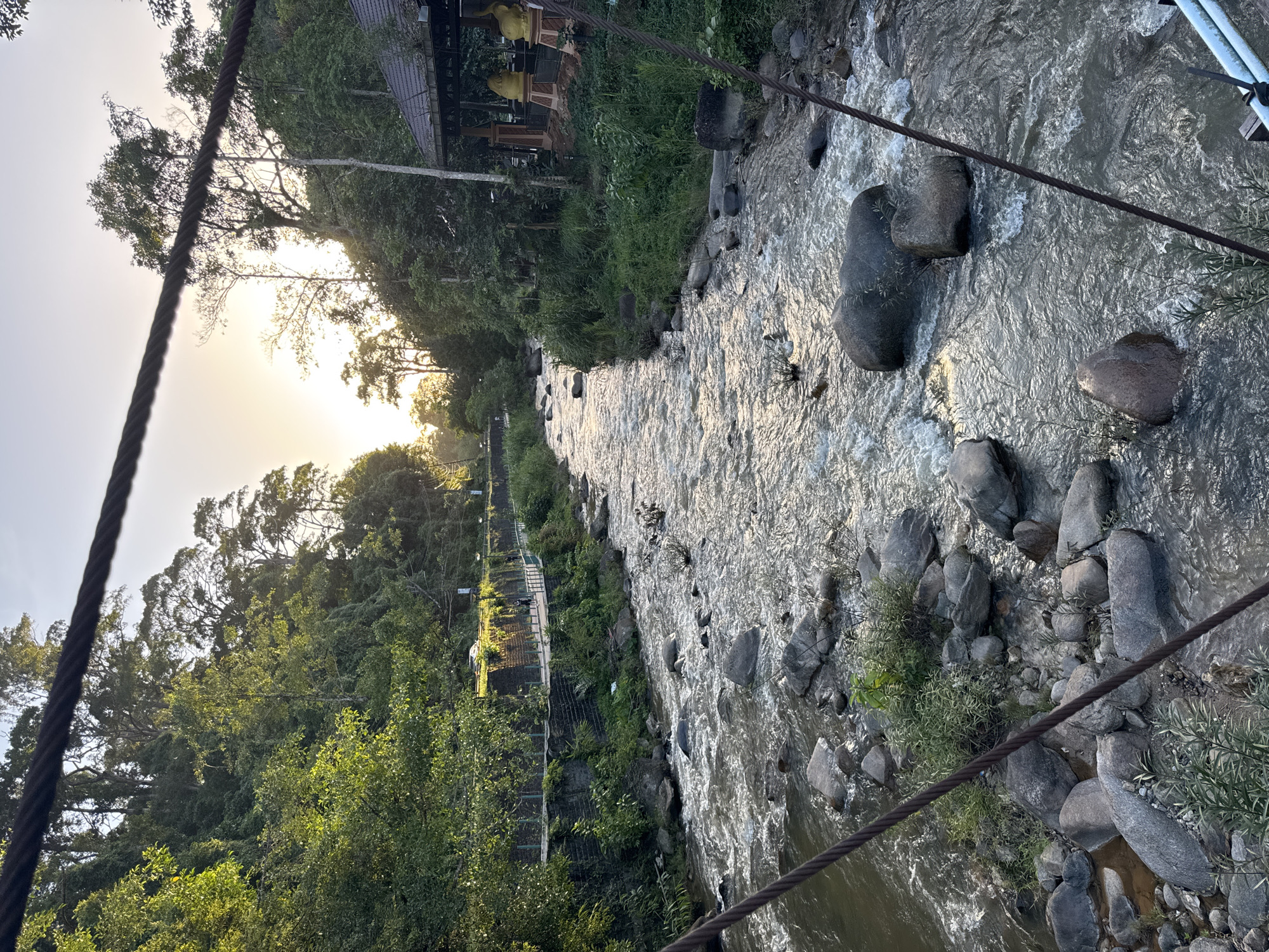 This is a photograph taken from a bridge overlooking a river flowing through a lush, green environment in Ranong, Thailand. The sun is setting, casting a warm glow over the scene. On the left bank of the river, a pathway with a stone wall and metal railings is visible, with trees and foliage growing above it. Further in the distance, a few people can be seen walking along the pathway. On the right bank, there's a covered structure with a tiled roof and decorative elements, possibly a resting area or a shrine, with two large golden statues visible. The river itself is wide and filled with numerous rocks of varying sizes, creating ripples and small rapids as the water flows. The surrounding vegetation is dense, with tall trees and undergrowth characterizing the landscape. The photograph is framed by thick cables in the foreground, indicating the perspective is from a suspension bridge.