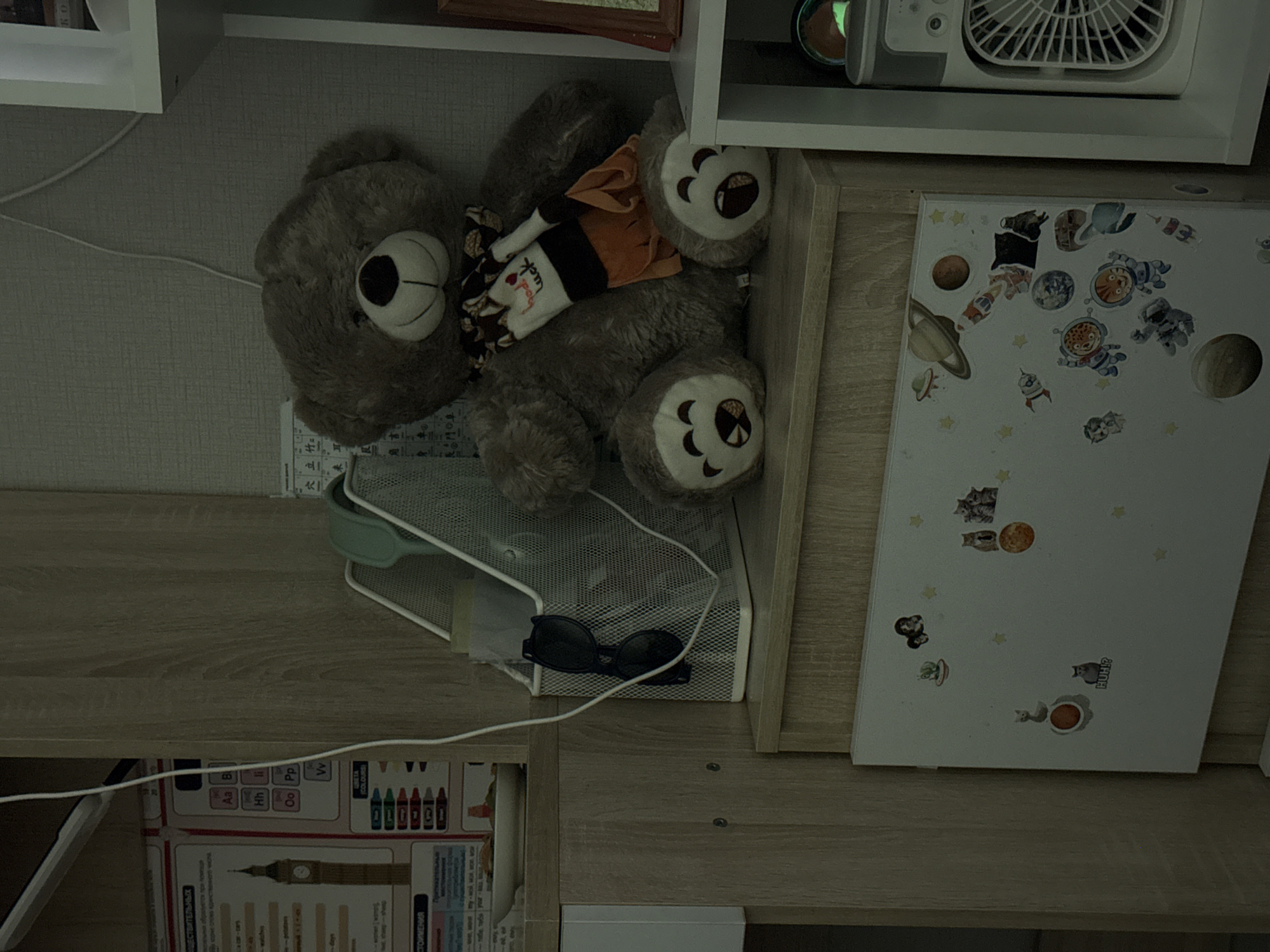 A dimly lit, indoor scene features a large, fluffy grey teddy bear with a patterned scarf around its neck, sitting on a wooden shelf. To the left of the bear, a white metal organizer holds a pair of black sunglasses. Behind the organizer, a poster with images of Big Ben and text in Russian is partially visible. Below the shelf, a white magnetic board is adorned with various stickers, including cats, planets, and astronauts. To the right of the teddy bear, a white shelf unit holds a small fan with a visible grille. The overall environment suggests a personal space, possibly a room in an apartment or home. The image does not provide any specific geographical or contextual clues to pinpoint its location in Cheboksary, Russia.