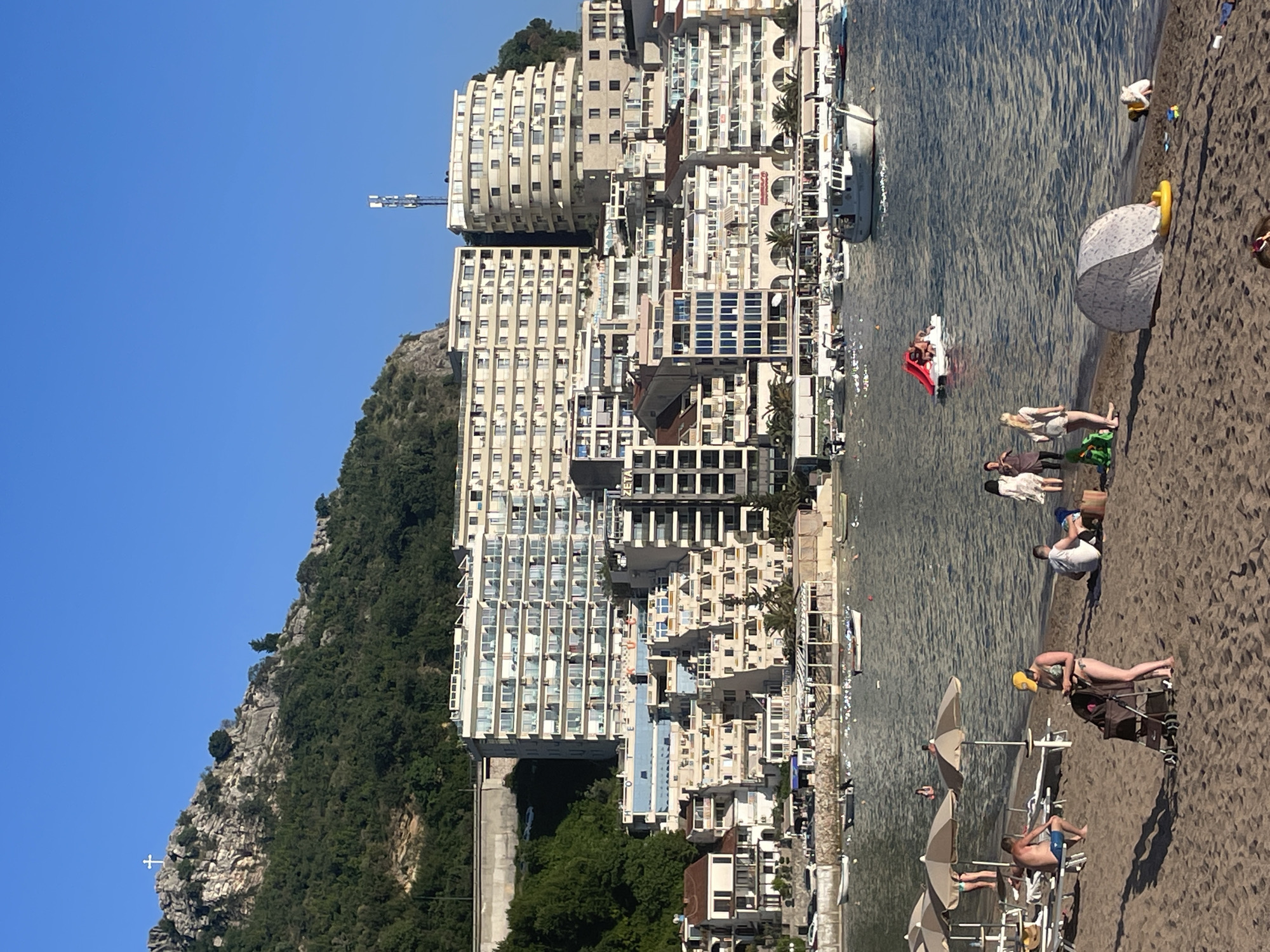 The image depicts a coastal scene in Rafailovići, Montenegro. Tall, multi-story residential buildings, likely apartments or hotels, line the shore. These buildings are light-colored and feature rows of balconies. The architecture appears modern. A mountain slope covered with greenery rises in the background. In the water, there is a boat with people, and the water's surface is slightly rippled. On the sandy beach, several people are present, some lying down, others sitting in chairs, and some are walking along the shoreline. Beach umbrellas are scattered along the beach. The sky is a clear, bright blue.