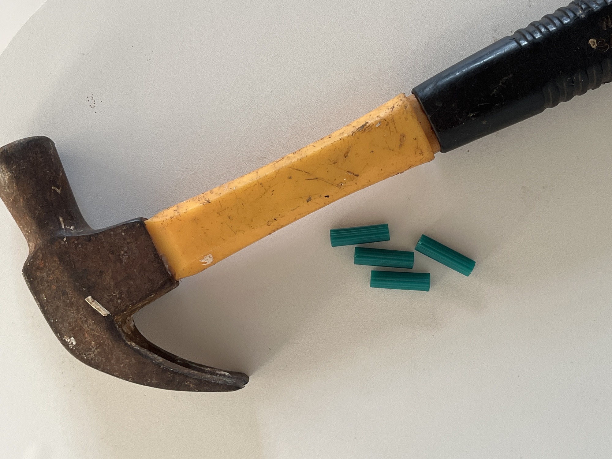 A close-up overhead shot shows a worn, metal claw hammer with a yellow handle and a black grip lying on a textured white surface. To the left of the hammer, four small, green cylindrical objects are scattered. The image is devoid of human presence or any discernible urban context, making it impossible to determine a specific location within Teluk Air, Indonesia.
