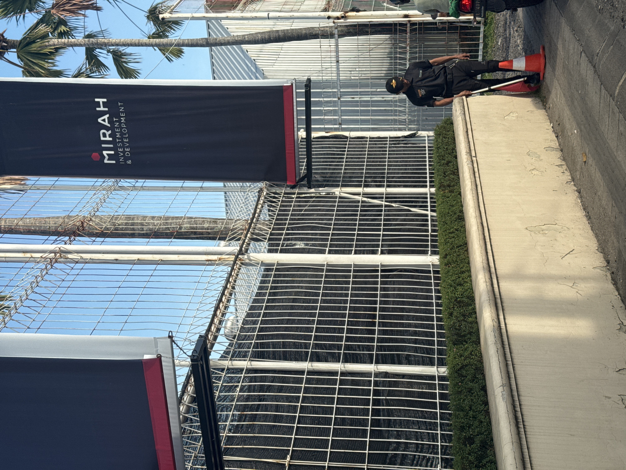 The image captures a construction site in what appears to be North Kuta, Indonesia, based on the "Mirah Investment & Development" banner displayed prominently on the fence. A security guard is standing along the sidewalk next to the construction fence. The construction site is enclosed by a tall, chain-link fence covered with black mesh, likely for privacy and dust control. A narrow strip of greenery separates the fence from the sidewalk. The sidewalk is concrete. The sky is visible through the gaps in the fence. A palm tree is also visible in the background. The security guard is in black uniform and holding what appears to be a traffic cone.