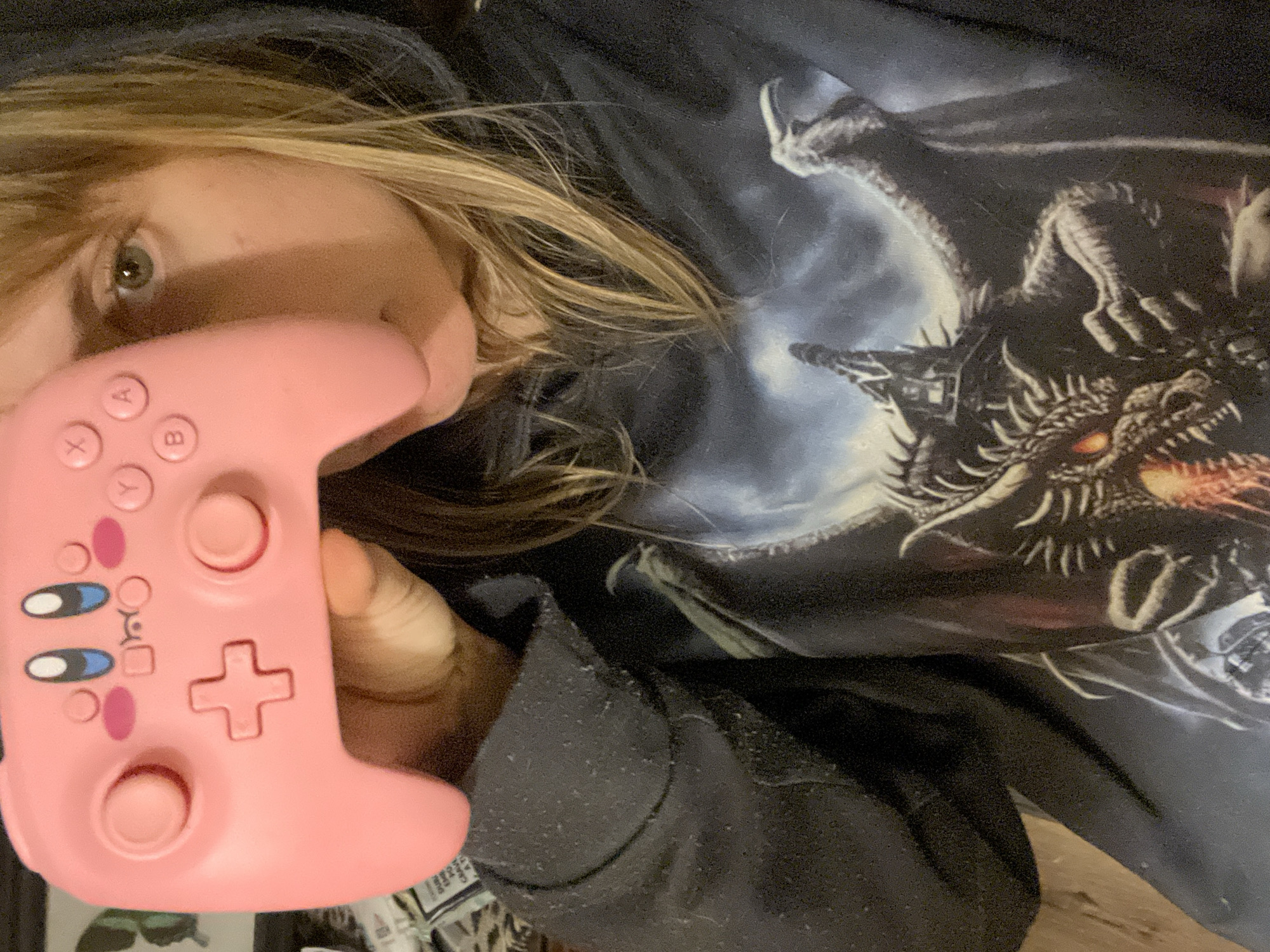 A person with blonde hair, fair skin, and green eyes is holding a pink video game controller. The controller has two joysticks, a directional pad, and buttons labeled X, Y, A, and B. The controller also has cartoonish eyes and blush marks on it. The person is wearing a dark-colored hooded sweatshirt. The shirt underneath the sweatshirt has a dragon printed on it. The background includes what seems to be a wooden floor. The location is not discernible from the image.