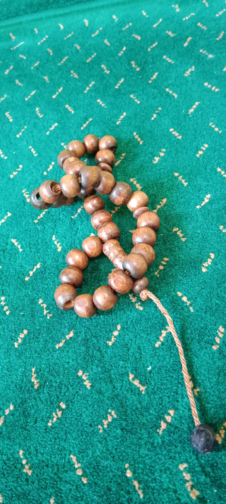 A close-up, overhead shot shows a rosary made of dark brown wooden beads on a green carpet with a faint tan pattern. The rosary is coiled loosely, with some beads overlapping. A thin, braided tan string extends from the rosary, ending in a single black bead. The background is a textured green carpet with a repeating diagonal pattern of small, light tan marks. The image appears to have been taken indoors, likely in a residential setting in Soko, Indonesia, given the context provided. There are no people or animals present in the image. The lighting suggests a natural light source, possibly from a window.
