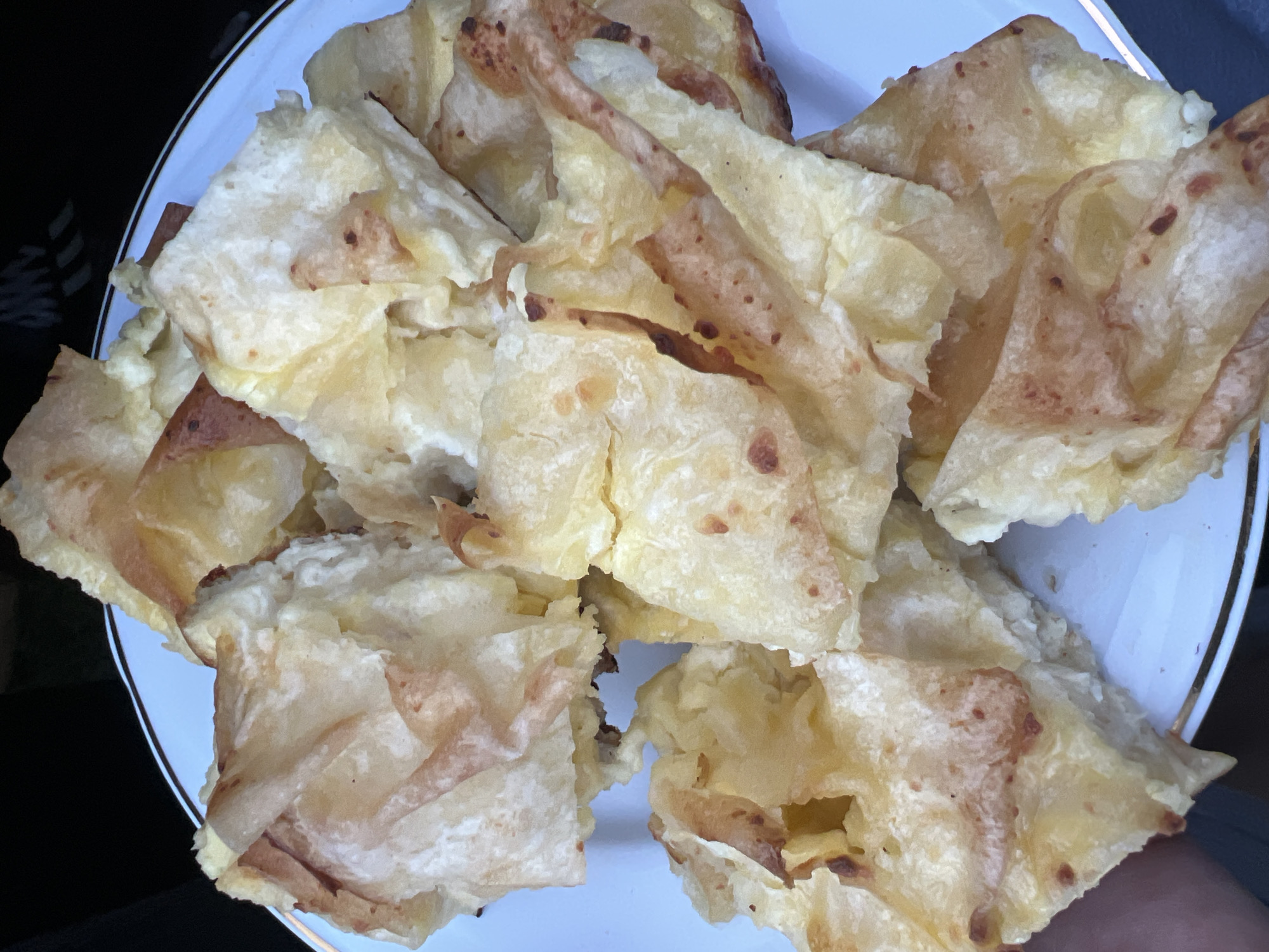 A close-up, overhead shot displays a white plate filled with pieces of golden-brown, layered pastry. The pastry appears to be flaky, with some pieces showing slightly darker, crisped edges and lighter, softer interiors. The pastry is cut into squares or rhomboid shapes, piled unevenly on the plate. The background is dark and out of focus, offering no discernible environmental details or location information. There are no people or actions depicted in the image. The scene is static and focused solely on the food item.