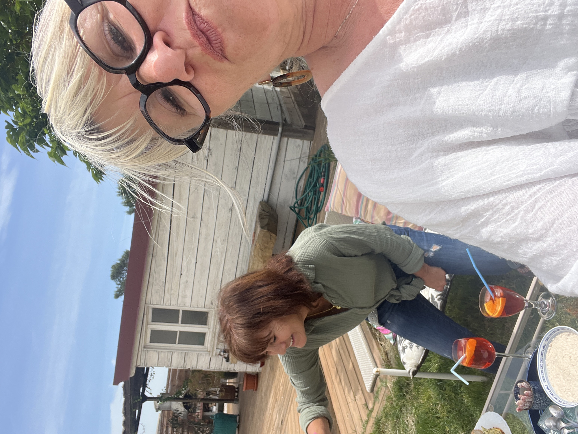 Two women are seated at an outdoor table in Montarnaud, France. One woman, seen close-up, has short blonde hair and wears glasses and a white blouse. The other woman has shoulder-length brown hair and wears a green button-down shirt and jeans. Two spritz cocktails with straws and orange slices sit on the glass table, along with plates of food. A white wooden outbuilding is visible in the background. The table sits on a lawn next to a wooden deck.