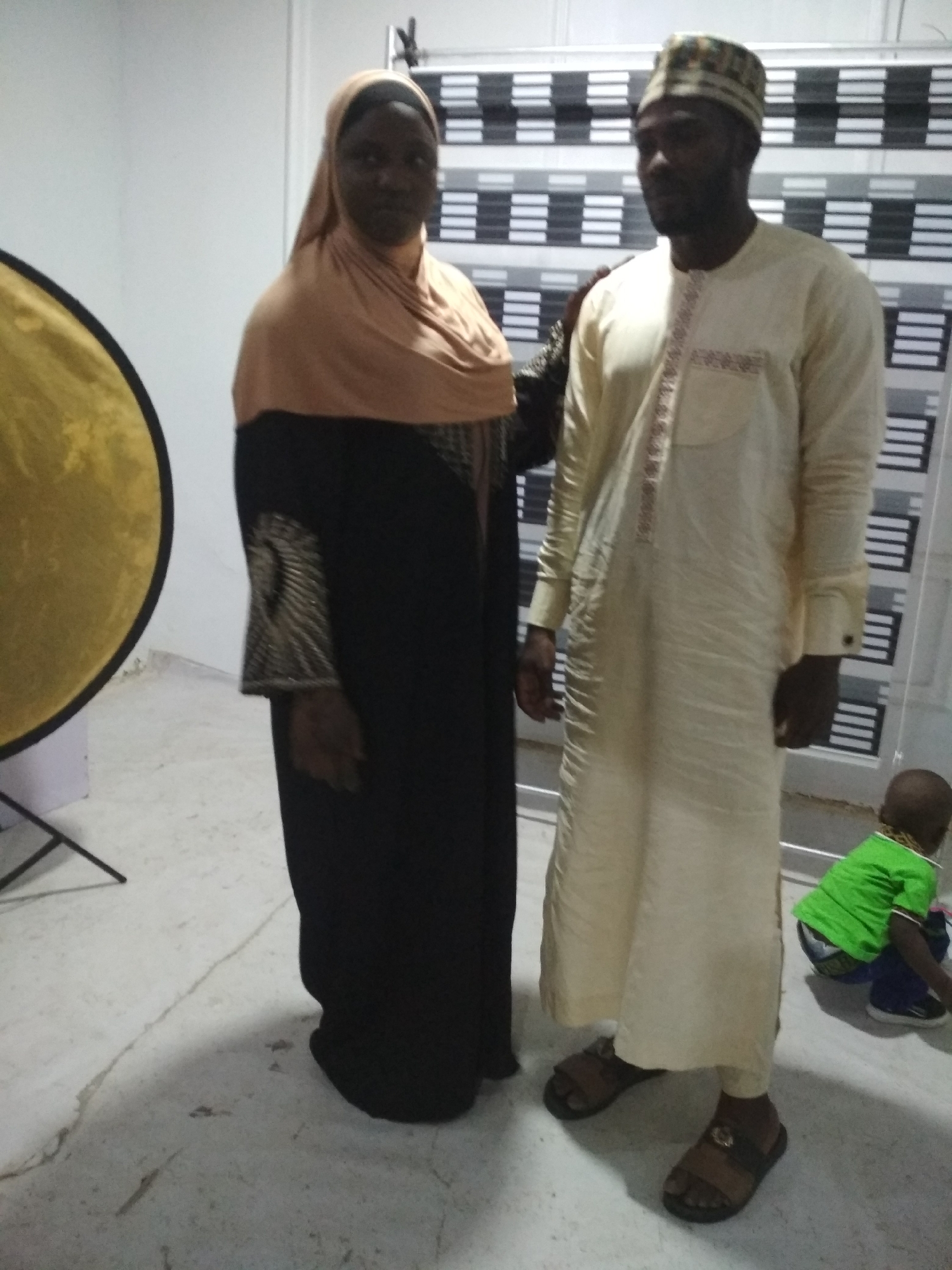 A medium, eye-level shot shows two adults standing together with a small child squatting in the background. The woman on the left is wearing a long, black dress with decorative gold embellishments on the sleeves, and a peach-colored headscarf. The man on the right is wearing a light yellow, long-sleeved garment with a patterned strip running down the front, and a small, patterned cap. Both adults are wearing sandals. Behind them, there is a backdrop of alternating black and white stripes. The small child in the background is wearing a bright green shirt. To the left, there is a gold circular reflector. The location is in an interior space in the city of Unknown, Unknown.