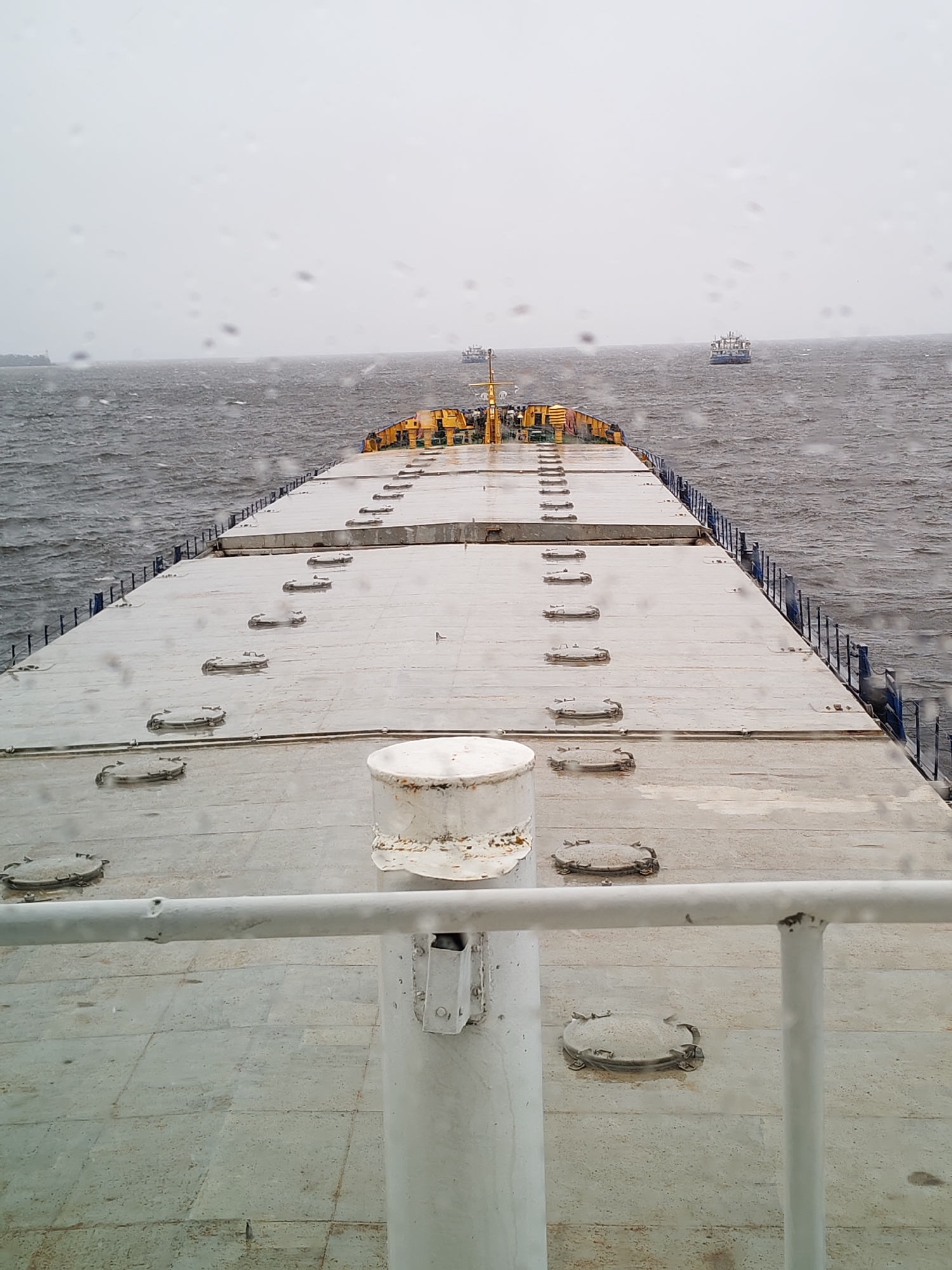 The image captures a stern view from a large cargo ship, looking forward over its extensive, flat deck. The deck, predominantly grey, features numerous oval-shaped cargo hatch covers arranged in rows, leading towards the ship's bow where some yellow-painted structures are visible. White railings line the sides of the deck, and a prominent white stanchion and railing are in the immediate foreground, showing signs of wear and rust.
The ship is underway on a body of water, likely a river or estuary, matching the context of Shcheleiki, Russia. The water is dark grey and choppy, indicating windy conditions. The sky is overcast and uniformly grey, contributing to an overall muted and somewhat somber atmosphere. Raindrops are clearly visible on what appears to be a window or camera lens in the foreground, suggesting a rainy or misty day.
In the distance, across the turbulent water, at least two other vessels are visible, appearing as dark shapes against the grey horizon. These distant ships are somewhat obscured by the weather and distance. No people are visible on any of the vessels. There is no discernible text in the image. The scene conveys a sense of rugged maritime travel in challenging weather conditions during daylight hours.