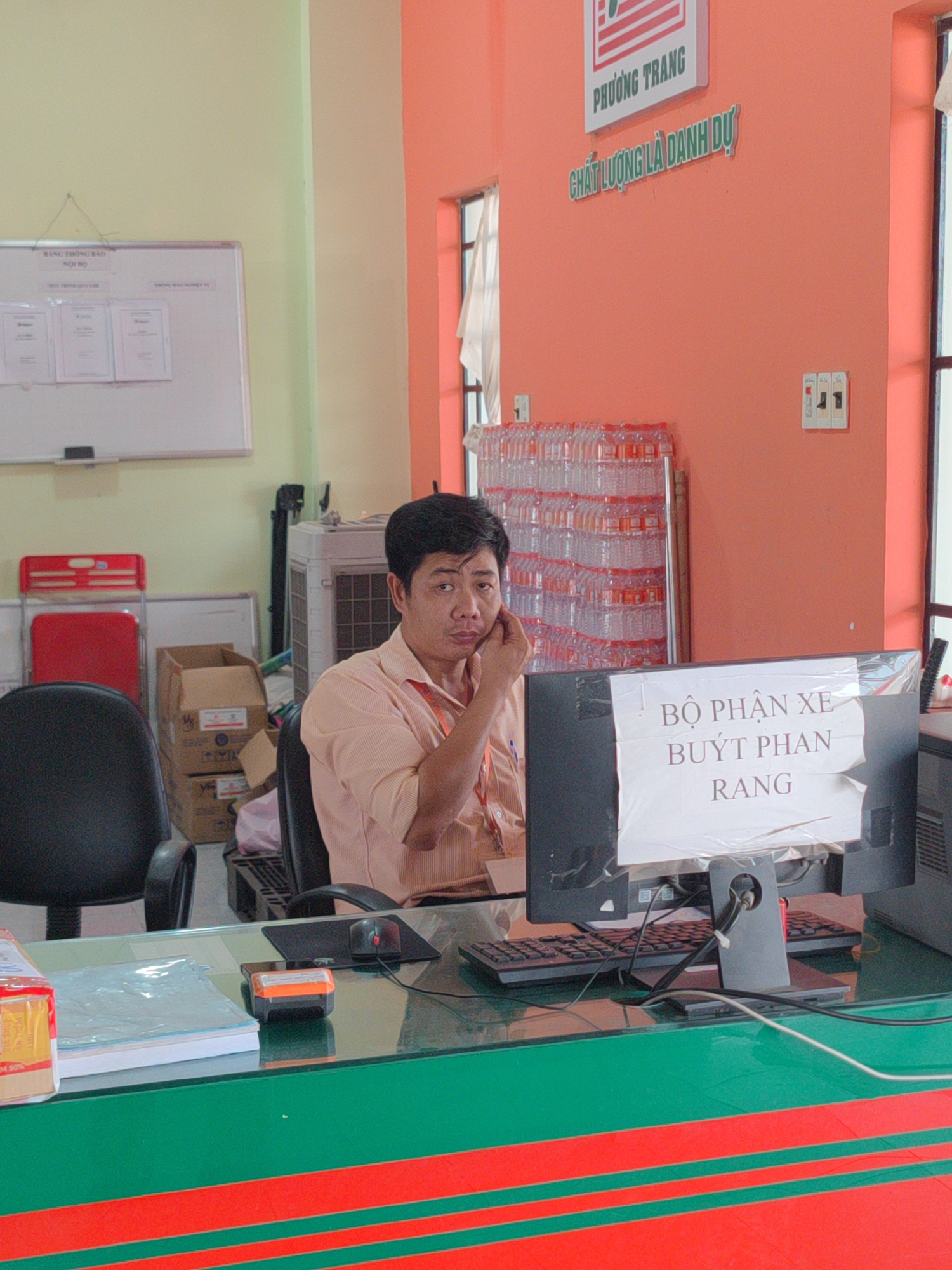 An indoor scene features a man seated at a desk, with a computer monitor in front of him. The screen has a sign taped to it that says "BỘ PHẬN XE BUÝT PHAN RANG". The man, who appears to be of Southeast Asian descent, is wearing a light orange shirt and is resting his chin on his hand, looking directly at the camera. The desk is covered in green and red stripes. Behind the man are a water cooler, a stack of bottled water, and a wall painted peach. A window with white curtains is partially visible in the background. Also visible are office items such as a white board with papers, a chair, a mouse, and a stapler.