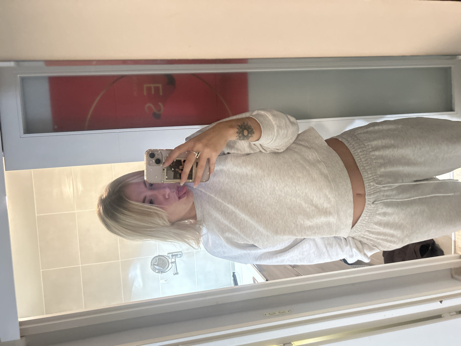 A fair-skinned woman with short blonde hair is taking a mirror selfie, sticking her tongue out slightly. She's wearing a light gray cropped sweatshirt and matching sweatpants, revealing a bit of her stomach. A circular tattoo is visible on her upper left arm. The mirror reflects a tiled wall and a window with a red design. The woman holds her phone in her right hand, showcasing a clear case with photos inside.