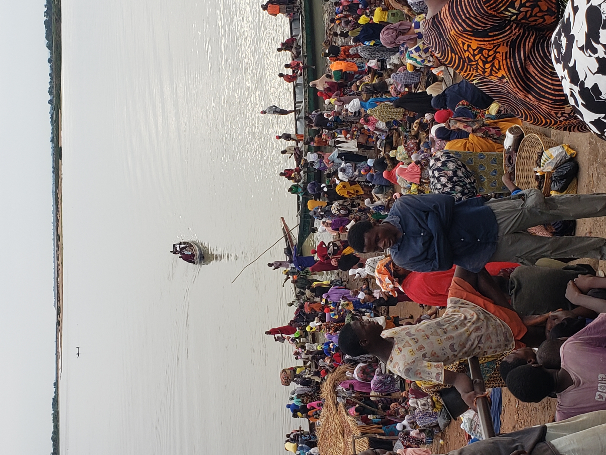 The image depicts a crowded waterfront scene in Lokoja, Nigeria. A large group of people, mostly wearing colorful clothing, are clustered along the shoreline and on a boat. The boat is positioned alongside the shore and filled with passengers. A smaller boat is visible on the water, further away from the shore. The water body stretches into the distance, with land visible on the opposite bank. The overall impression is one of a busy transportation hub or market area near a river or lake in Lokoja.