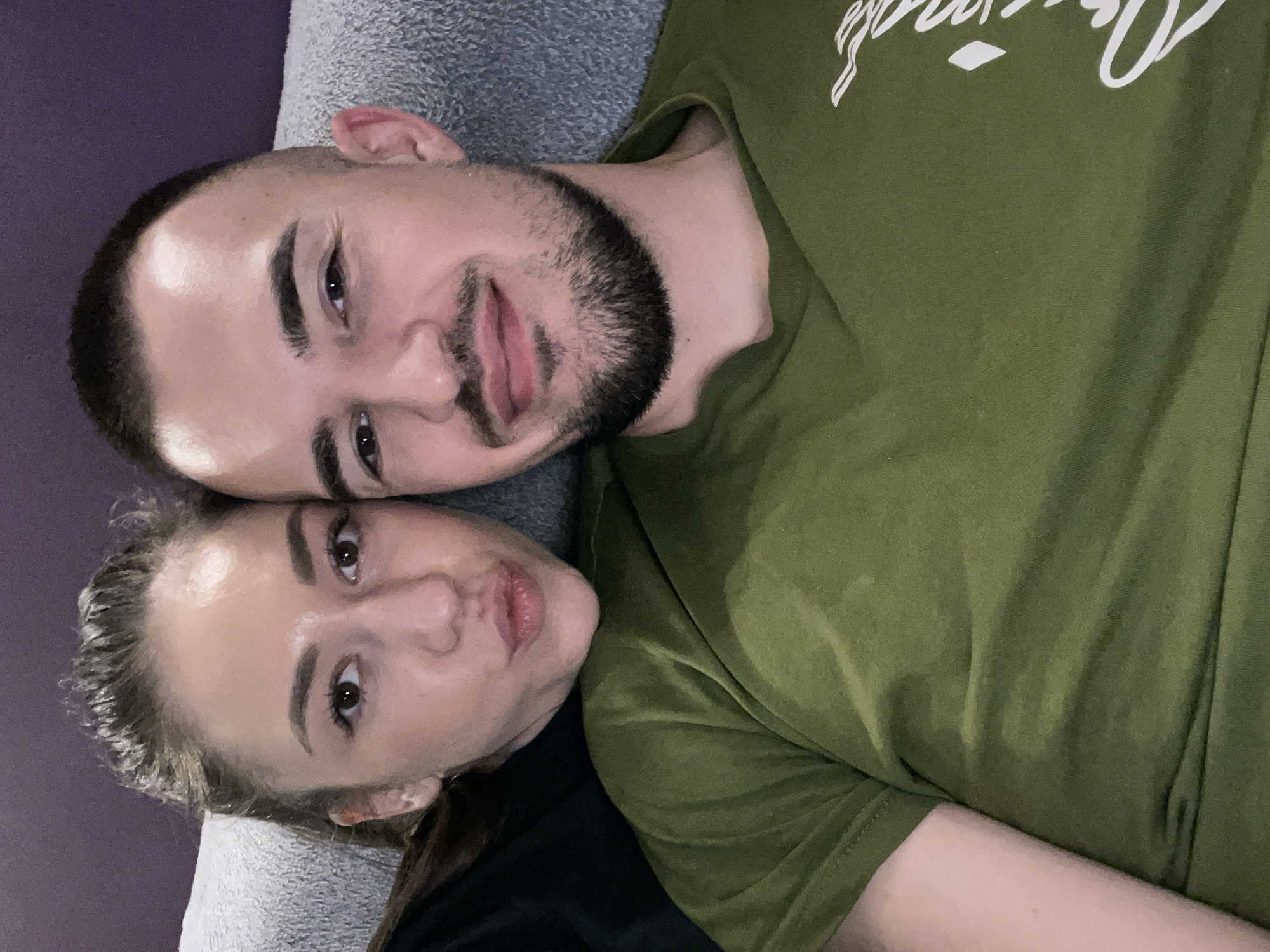 A couple is lying down. The man has short dark hair, a beard, and is wearing a green t-shirt with white writing. The woman has her hair pulled back and is wearing a black t-shirt. They are lying on a grey surface. Location: Sofia, Bulgaria is not discernible from the image.