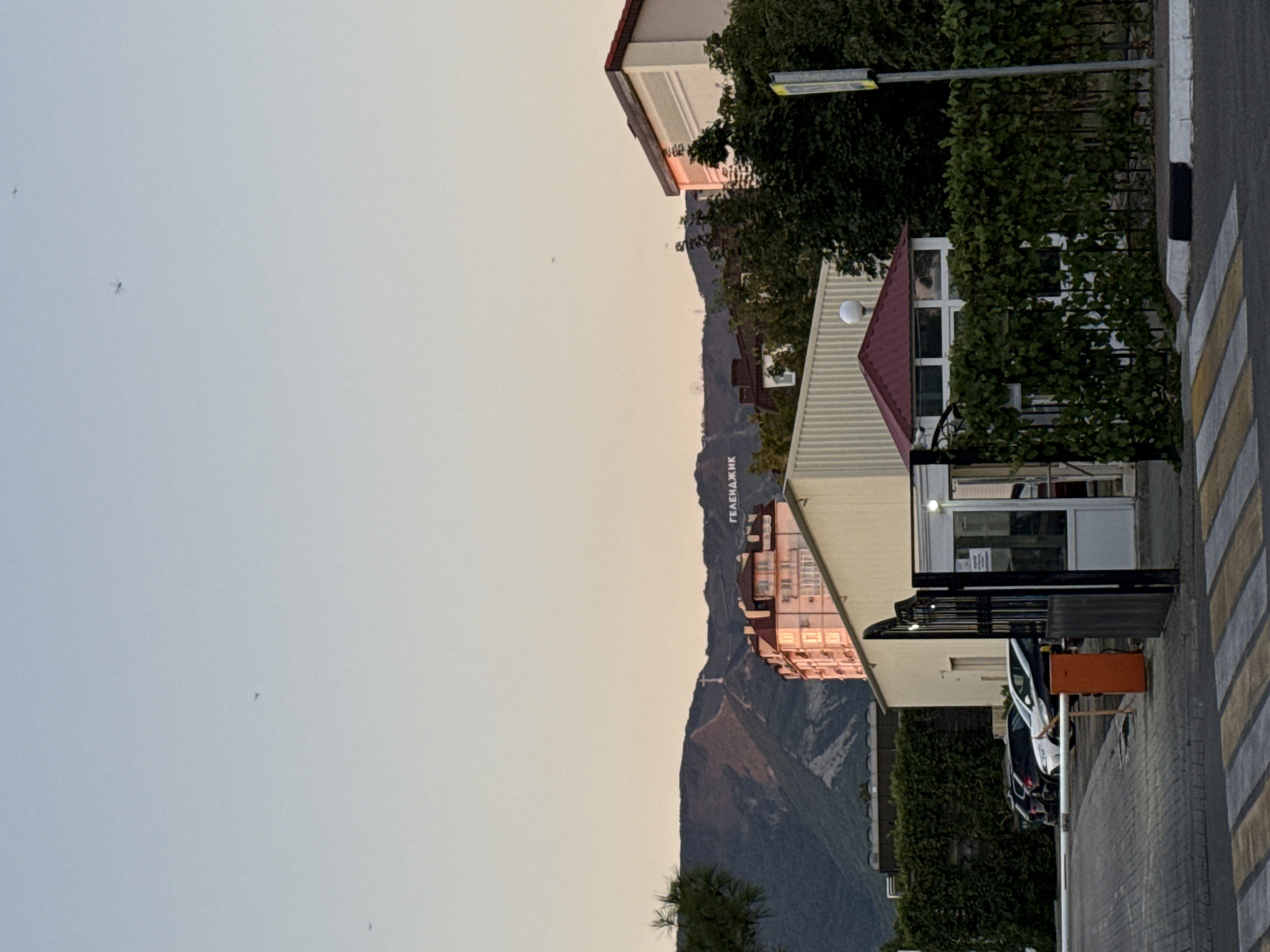 The image captures a panoramic view of the city of Gelendzhik, Russia, during what appears to be a late afternoon or early evening. The sky is a soft gradient of light blue and pale orange, indicative of sunset. In the distance, a prominent mountain range serves as a dramatic backdrop. A white, illuminated sign spelling out "ГЕЛЕНДЖИК" (Gelendzhik) is clearly visible on the slope of one of the mountains. To the right of the sign, a large statue of Jesus Christ stands atop a peak.
In the foreground, a gated entrance with black metal bars leads into a complex. Adjacent to the entrance is a small, white guardhouse with a red roof, partially obscured by lush green ivy growing on a fence. Several cars are parked in a lot behind the gate, including a white SUV, a dark SUV, and a black sedan. A pedestrian crossing with yellow and white stripes marks the immediate foreground. Behind the gate and guardhouse, a multi-story building with a pale yellow facade and red-tiled roof is visible, catching the warm glow of the setting sun. Further back, another taller building, also with illuminated windows, stands out. Trees and foliage are scattered throughout the scene, adding to the urban landscape. The overall impression is one of a peaceful, established urban area nestled against a natural mountainous terrain.