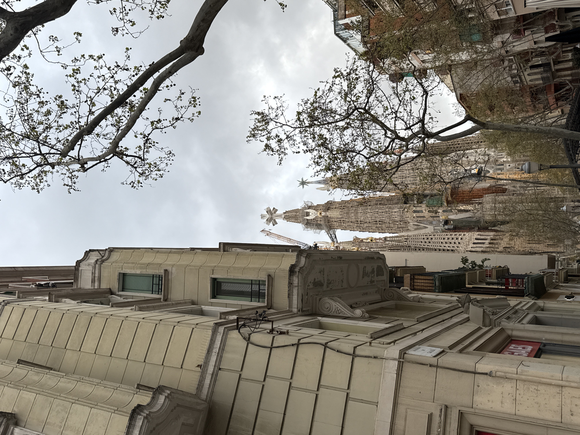 The image depicts an urban street scene in Barcelona, Spain, with the Sagrada Familia basilica as the prominent background subject.
In the foreground on the left, a multi-story building with a light beige or yellow facade occupies the left third of the frame. This building features horizontal decorative banding, ornate cornices, and balconies with dark railings. Windows are visible, some equipped with closed dark green roller shutters or blinds. Exposed black electrical conduit or wiring runs vertically along the building's side. A partial red sign with white lettering, appearing to read "GAES," is visible at street level on this building.
Dominating the central background is the Sagrada Familia, an intricate and monumental basilica under construction. Its multiple highly detailed spires, adorned with crosses and stars, rise towards the overcast sky. Prominent yellow and white construction cranes are visible atop and adjacent to the basilica's structure, indicating ongoing work. Some sections display scaffolding or green construction netting.
Scattered throughout the midground and right side of the image are several deciduous trees with relatively bare branches, showing sparse green leaves and characteristic seed balls, consistent with Plane trees. These trees partially obscure views of the Sagrada Familia and other multi-story buildings in the background, which vary in color and style, some featuring small balconies. A dark decorative streetlight fixture is partially visible in the lower right, beneath the trees. The sky is uniformly overcast with a light grey cloud cover. No individuals or specific actions are discernible within the frame.