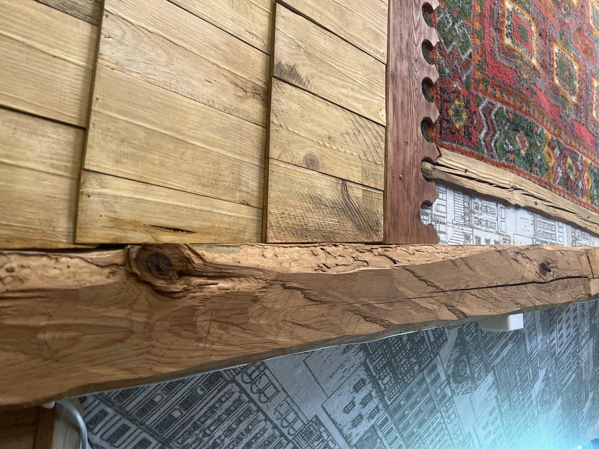An interior scene is depicted with a focus on architectural elements and decor. A large, rough-hewn wooden beam runs diagonally from the lower left to the upper right of the frame. To the left of the beam, a wall is visible, covered in wallpaper with a repeating pattern of architectural drawings, possibly depicting buildings or cityscapes. To the right of the beam, there are wooden planks forming a ceiling or wall. Below these planks, a decorative wooden trim with scalloped edges is attached, and beneath that, a colorful, intricately patterned rug is partially visible, hanging from the wall or ceiling. The lighting suggests an indoor environment, possibly natural light filtering in. The image appears to be taken from a low angle looking upwards. The location is identified as Daulėnai, Lithuania, implying this is likely a residential or historical interior. There are no people or actions visible in the image.