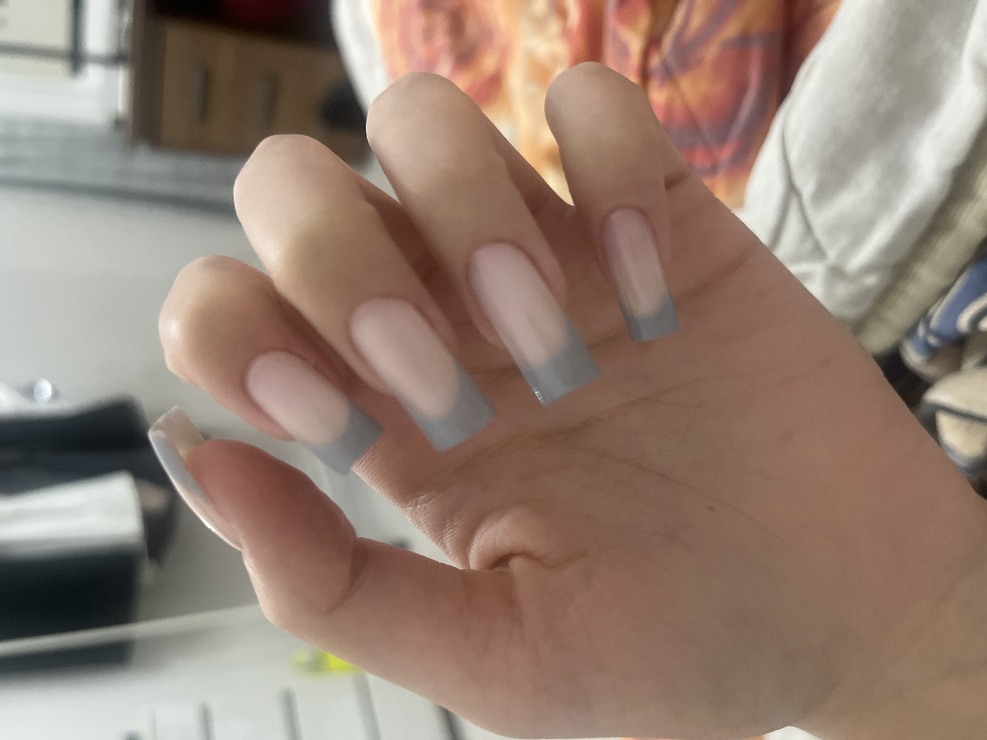 A close-up shot shows a person's hand with light pink and gray French tip acrylic nails. The hand is held against a blurry background of what appears to be a bedroom, with a bed, a wooden dresser, and white closet doors visible. The image quality is moderate, with some graininess and a shallow depth of field. The lighting is soft and diffused. There is no indication of the city of Perm, Russia, or any specific location within it.