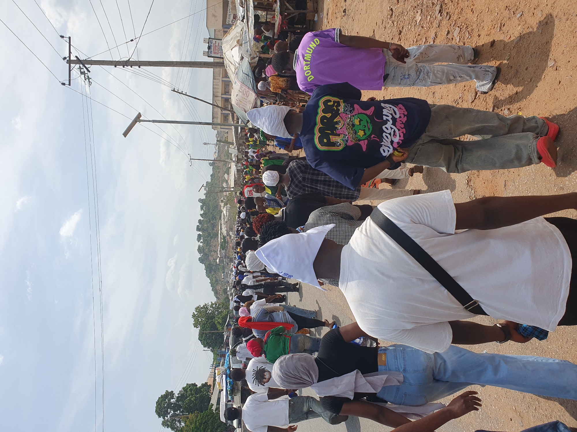 A large group of people are walking down a street in Lokoja, Nigeria. Most are wearing white head coverings. The street is paved, and there are buildings and power lines on the sides of the street. The people appear to be participating in a procession or march.